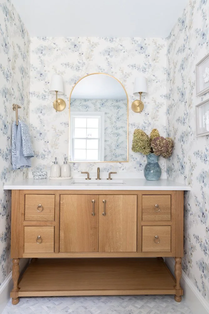 Bright bathroom with floral wallpaper, arched gold-framed mirror, brass fixtures, and wooden double vanity.
