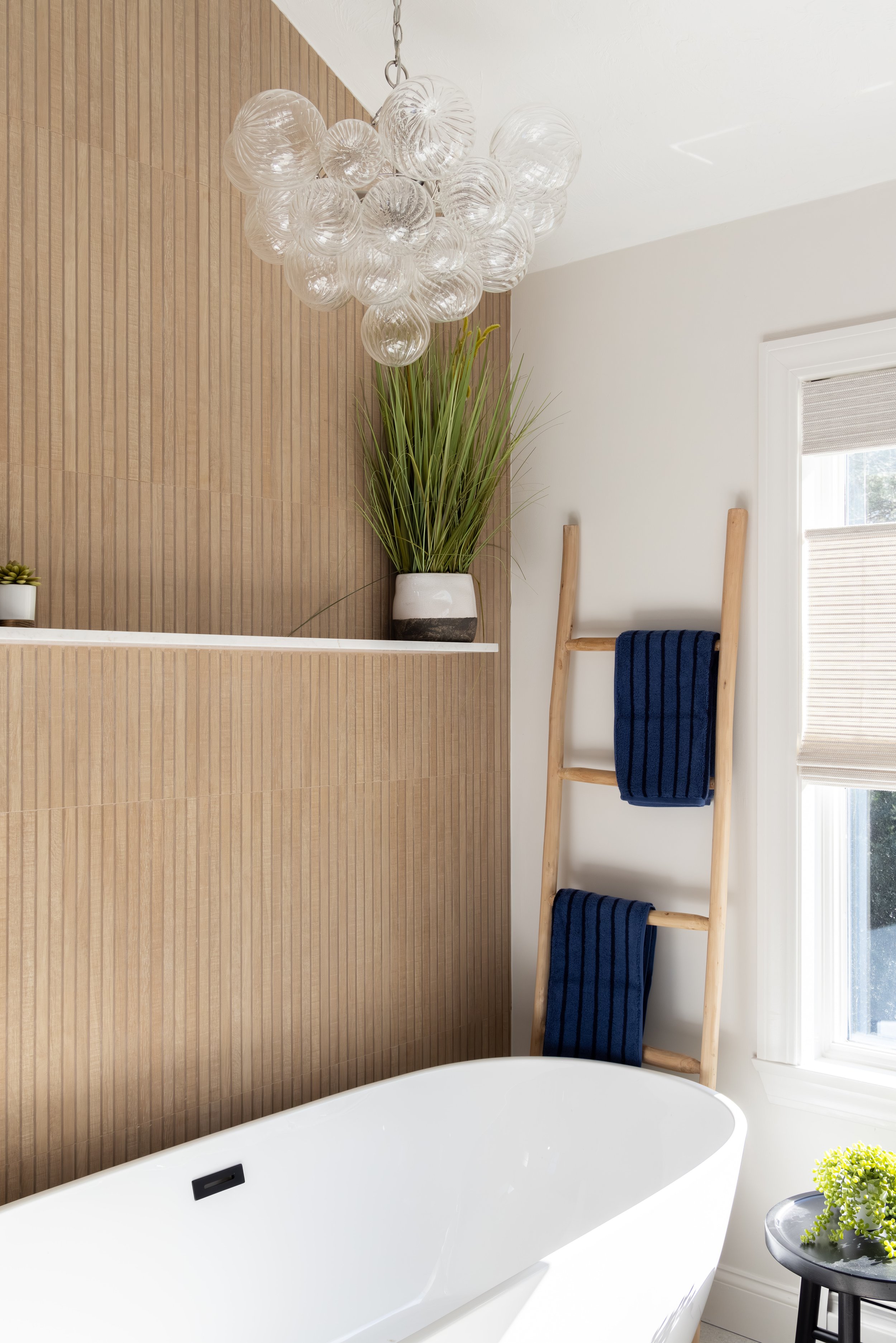 Modern bathroom with a freestanding white bathtub, glass globe chandelier, wood-paneled wall, ladder towel rack, and potted plants.