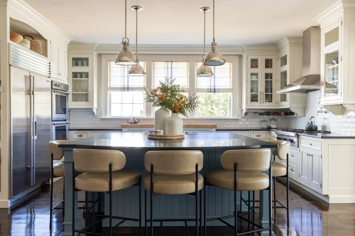 Bright white kitchen with a large dark island, tan bar stools, pendant lights, stainless steel appliances, and floral greenery centerpiece