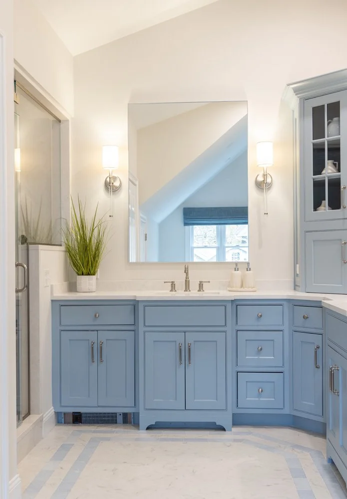 Bright bathroom vanity with blue cabinetry, white countertop, large mirror, wall sconces, and potted grass plant.