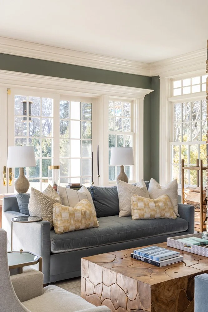 Bright traditional living room with a gray sofa, patterned pillows, large white-framed windows, table lamps, and a natural wood coffee table.