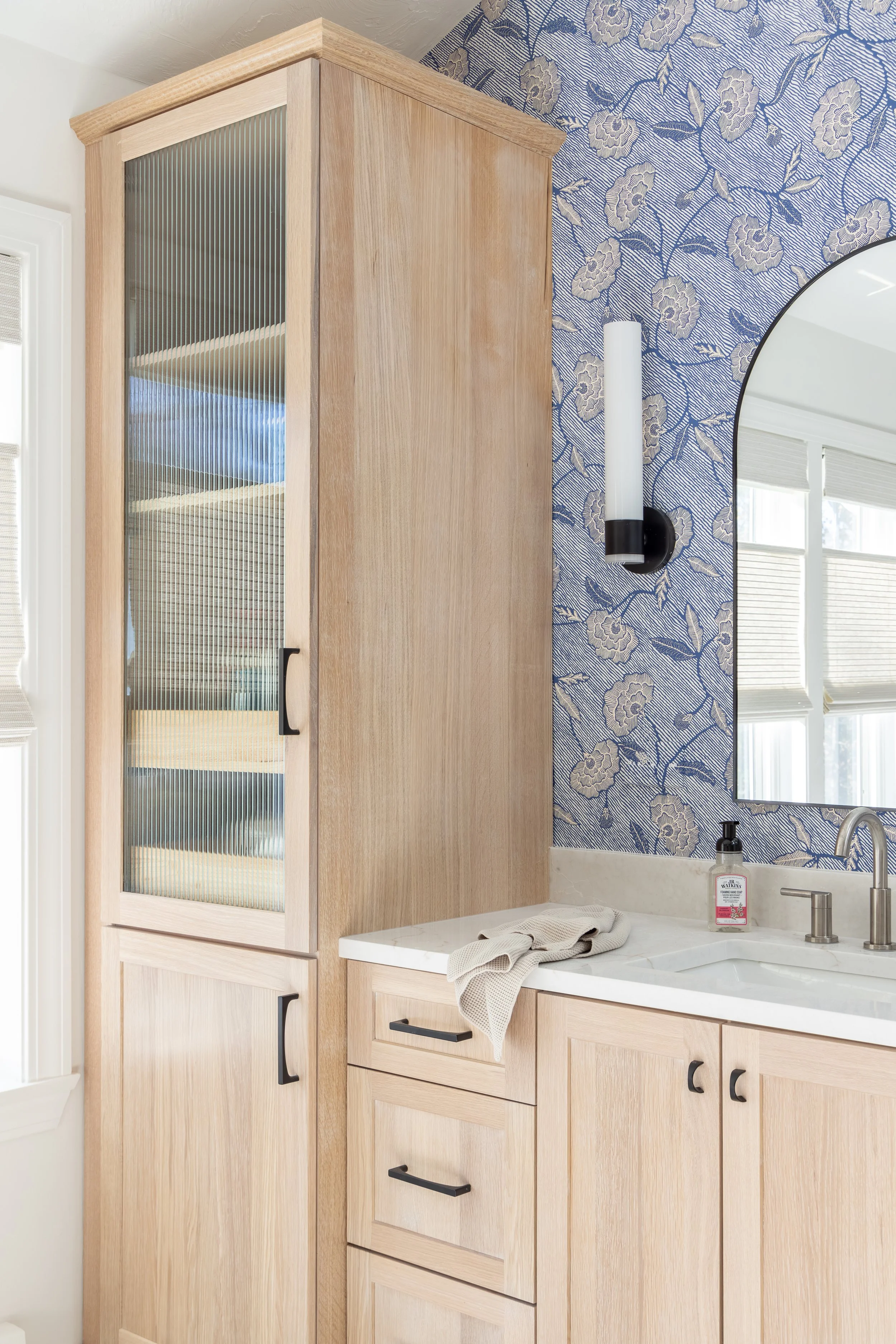 Modern bathroom vanity with light wood cabinetry, fluted glass door, white countertop, black fixtures, floral wallpaper, and arched mirror