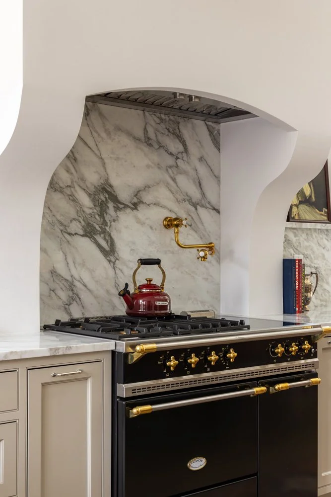 Luxury kitchen range with black and brass stove, red kettle, marble backsplash, and sculptural white hood surround