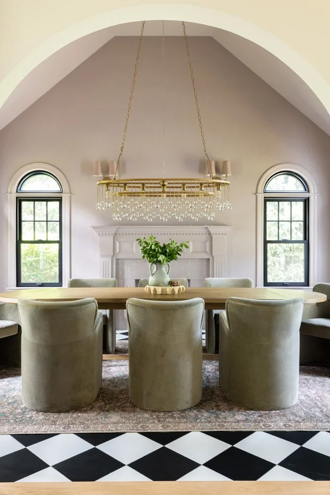 Elegant dining room with a long wooden table, upholstered chairs, crystal chandelier, fireplace, arched windows, and black-and-white checkerboard floor