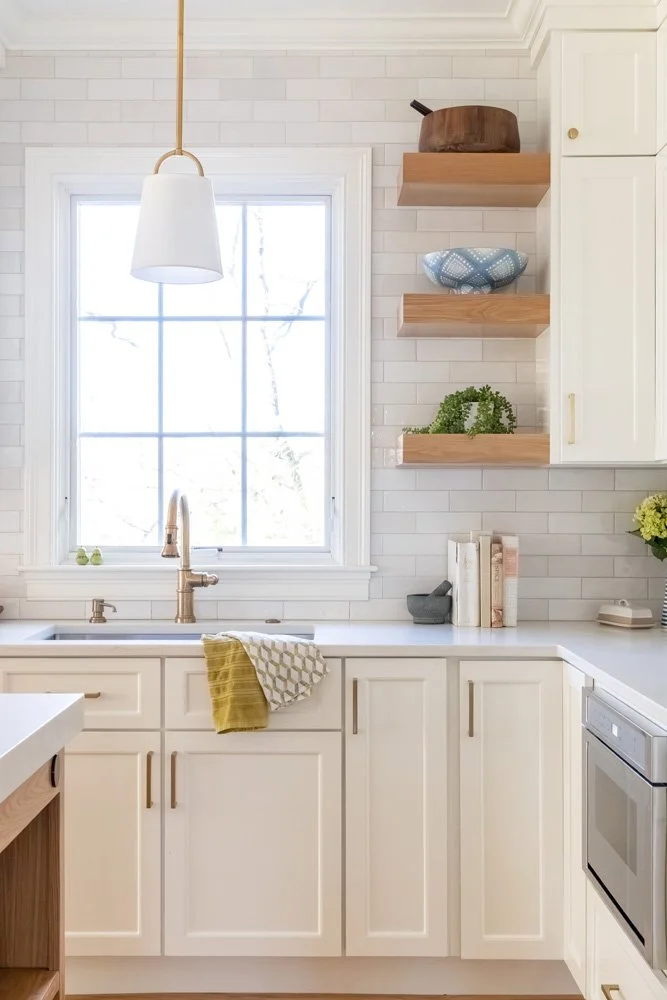 Bright white kitchen with farmhouse sink, brass pendant light, open wood shelves, subway tile backsplash, and stainless steel dishwasher
