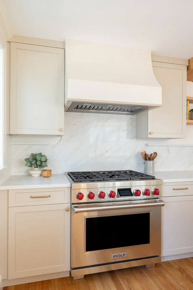 Bright modern kitchen with a stainless steel Wolf gas range, white cabinets, marble backsplash, and wood flooring.