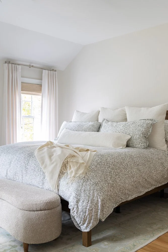 Bright white bedroom with a neatly made bed, patterned gray comforter, layered pillows, cream throw blanket, upholstered bench, and window with curtains