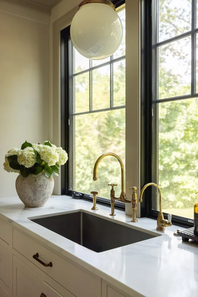 Elegant kitchen sink with brass faucets, white countertop, hydrangea bouquet, and large windows