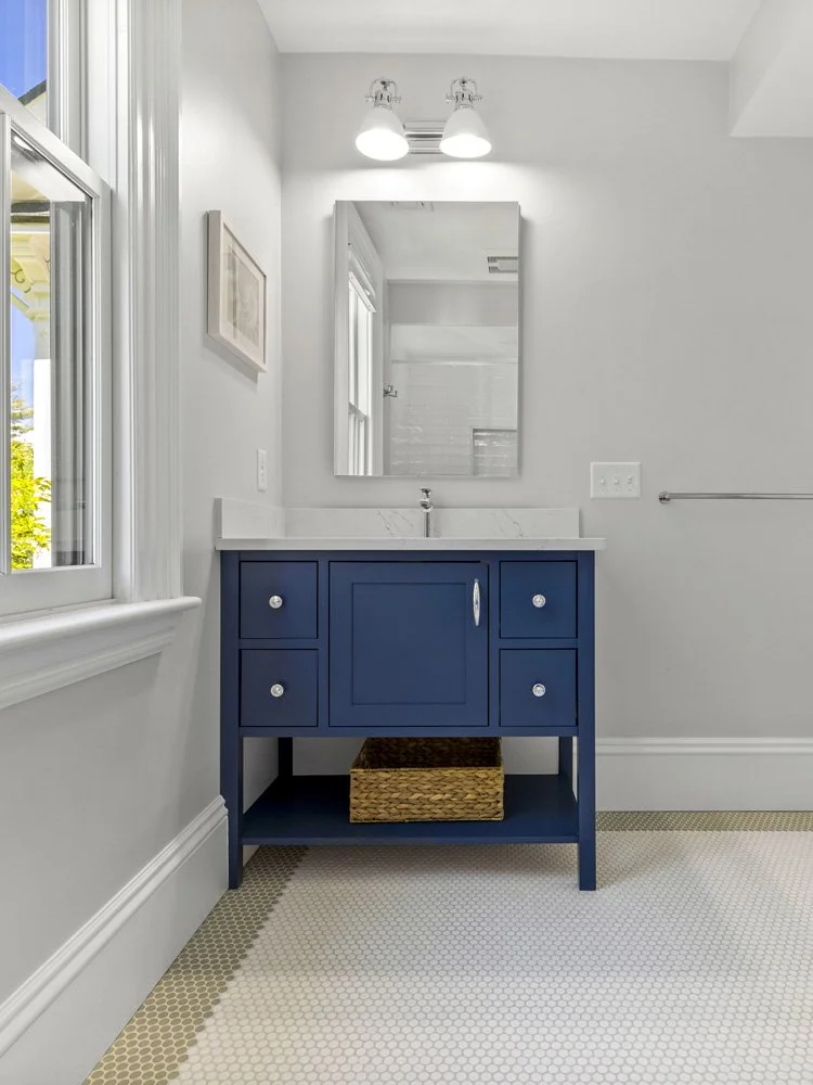 Bright bathroom with a navy blue vanity, marble countertop, rectangular mirror, double vanity lights, woven basket, and white hexagonal tile floor.