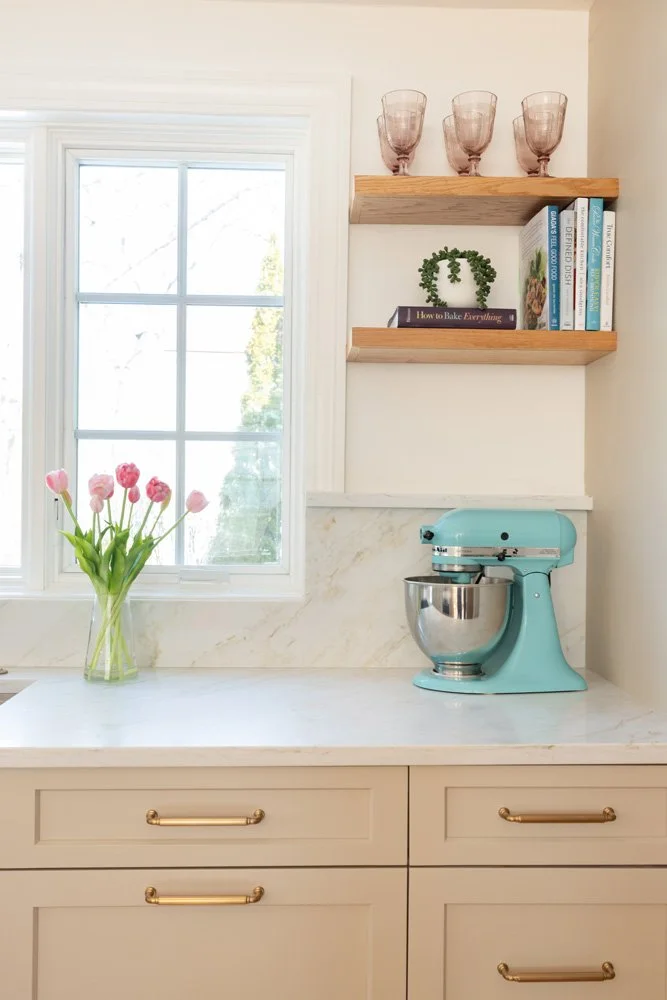 Bright kitchen corner with turquoise stand mixer, pink tulips, marble countertop, window, and wooden shelves with books and glasses
