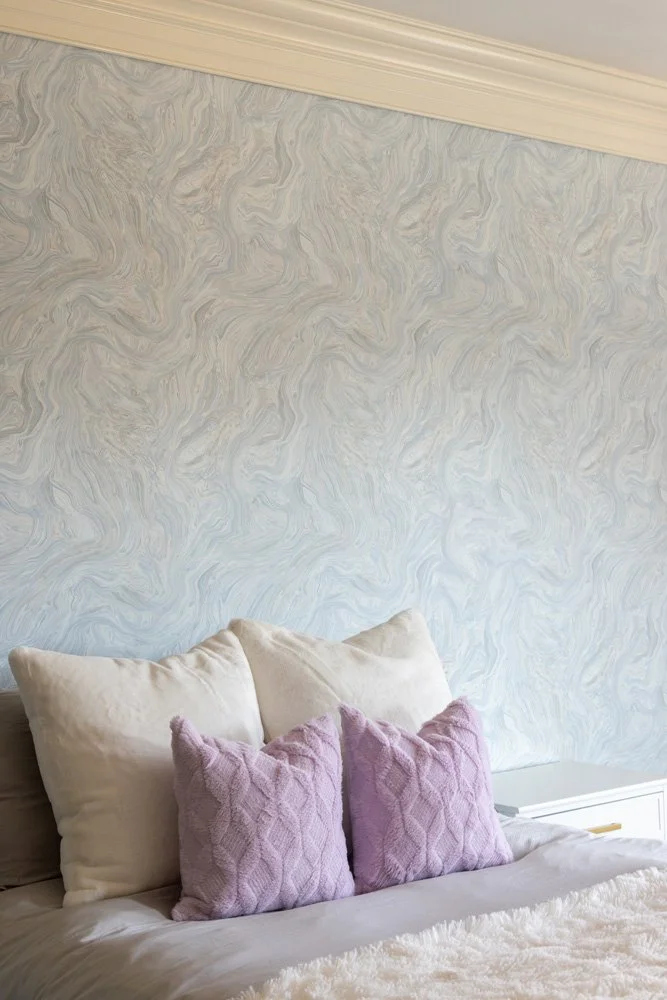 Cozy bed with white and lavender pillows against a pale blue marbled wallpapered wall
