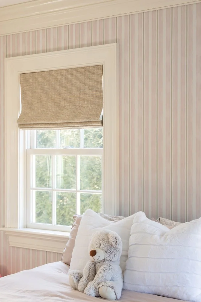 Cozy bedroom with pink striped wallpaper, a woven window shade, white bedding, and a plush teddy bear