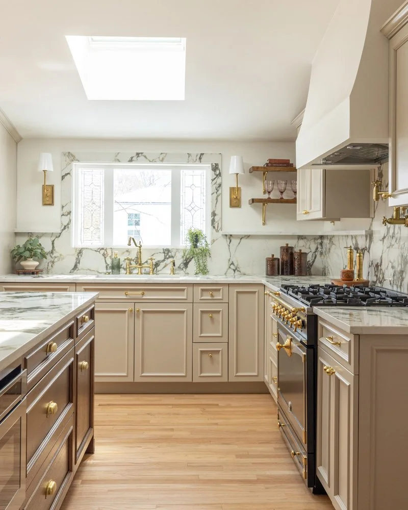 Elegant cream kitchen with marble countertops and backsplash, brass fixtures, central island, gas range, open shelving, and a sunlit window