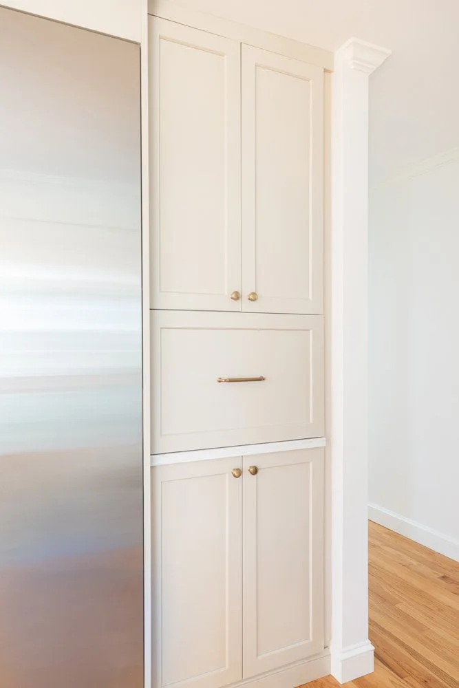Built-in white cabinetry with paneled doors, brass knobs, and a center drawer beside a stainless steel refrigerator.