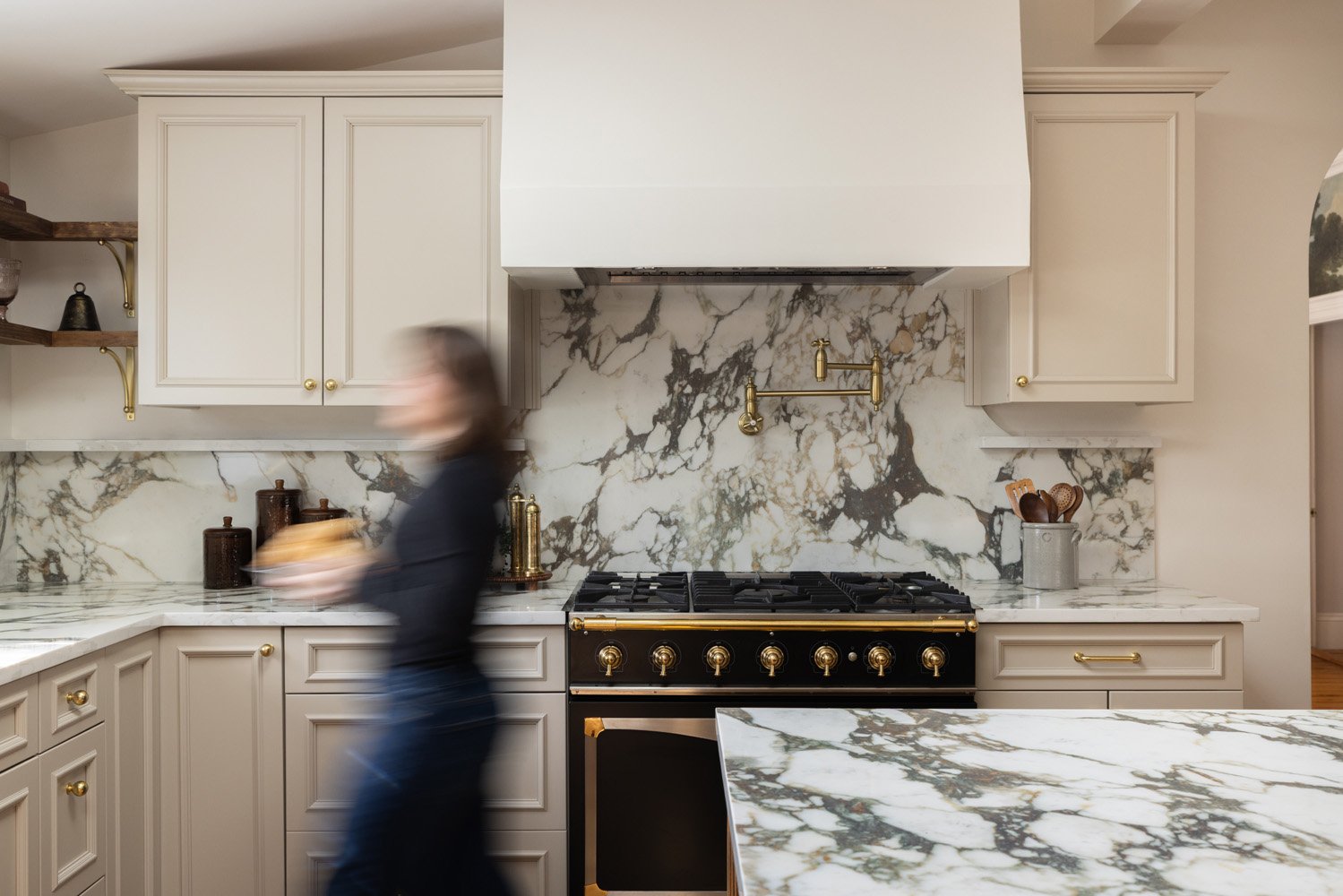 Elegant cream-colored kitchen with marble countertops and backsplash, brass fixtures, a black-and-gold range, and a blurred person walking in front