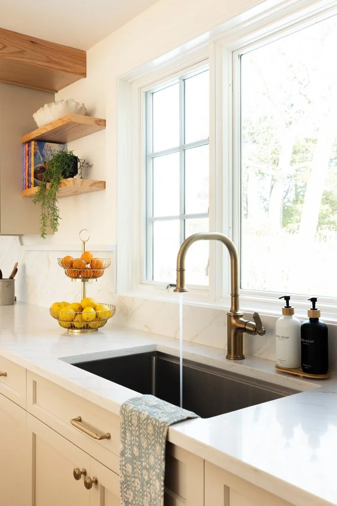 Bright white kitchen with farmhouse sink, brushed brass faucet, fruit baskets, open wood shelves, and large windows