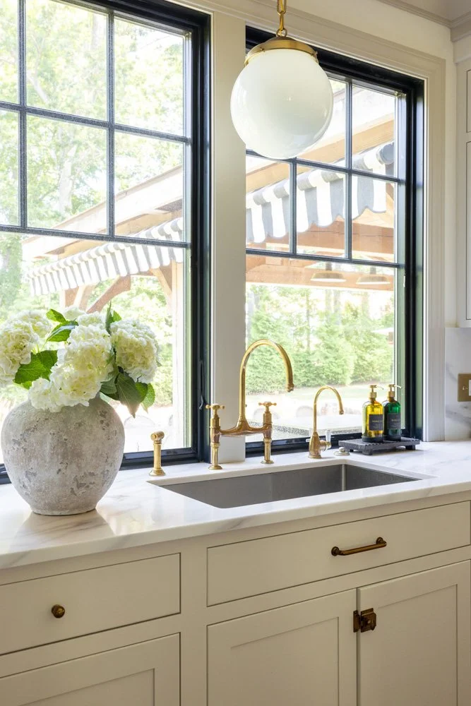 Bright kitchen sink with brass faucet, white quartz countertop, hydrangeas, and large black-framed windows