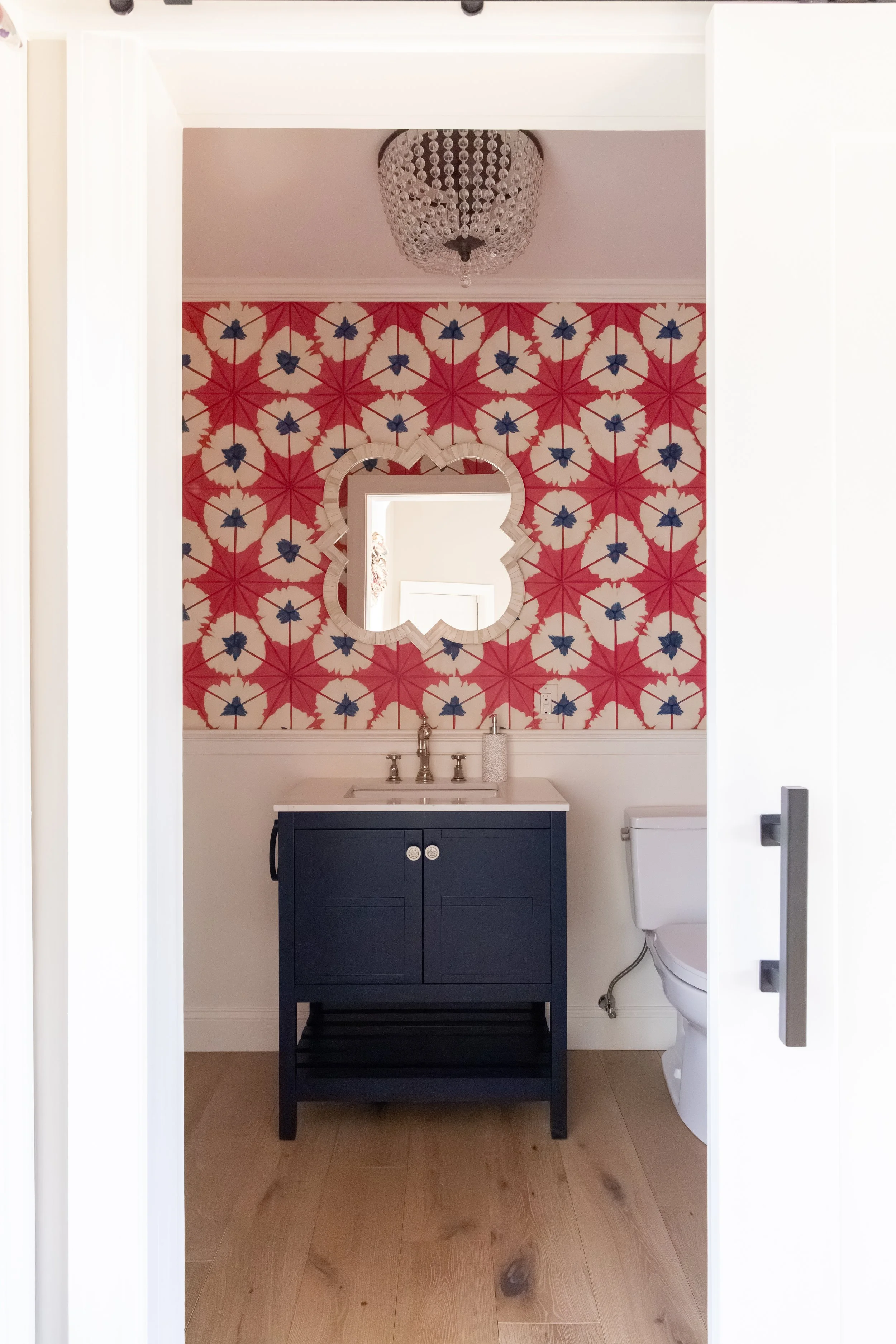 Small bathroom with red floral wallpaper, crystal chandelier, heart-shaped mirror, navy vanity, and white toilet