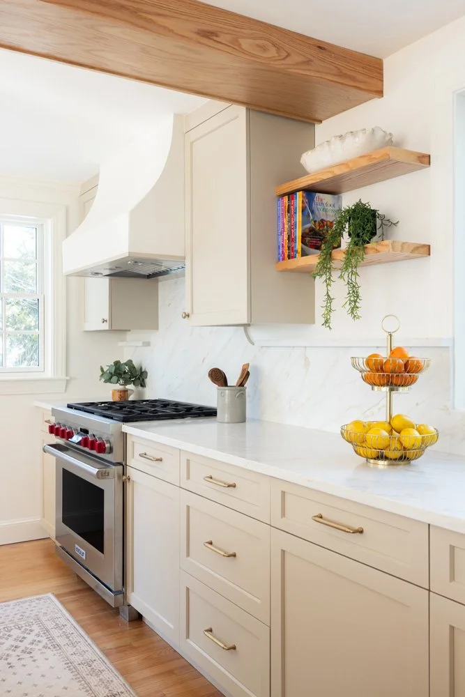 Bright white kitchen with marble countertops, stainless steel range, open wood shelves, potted herbs, and a tiered fruit basket