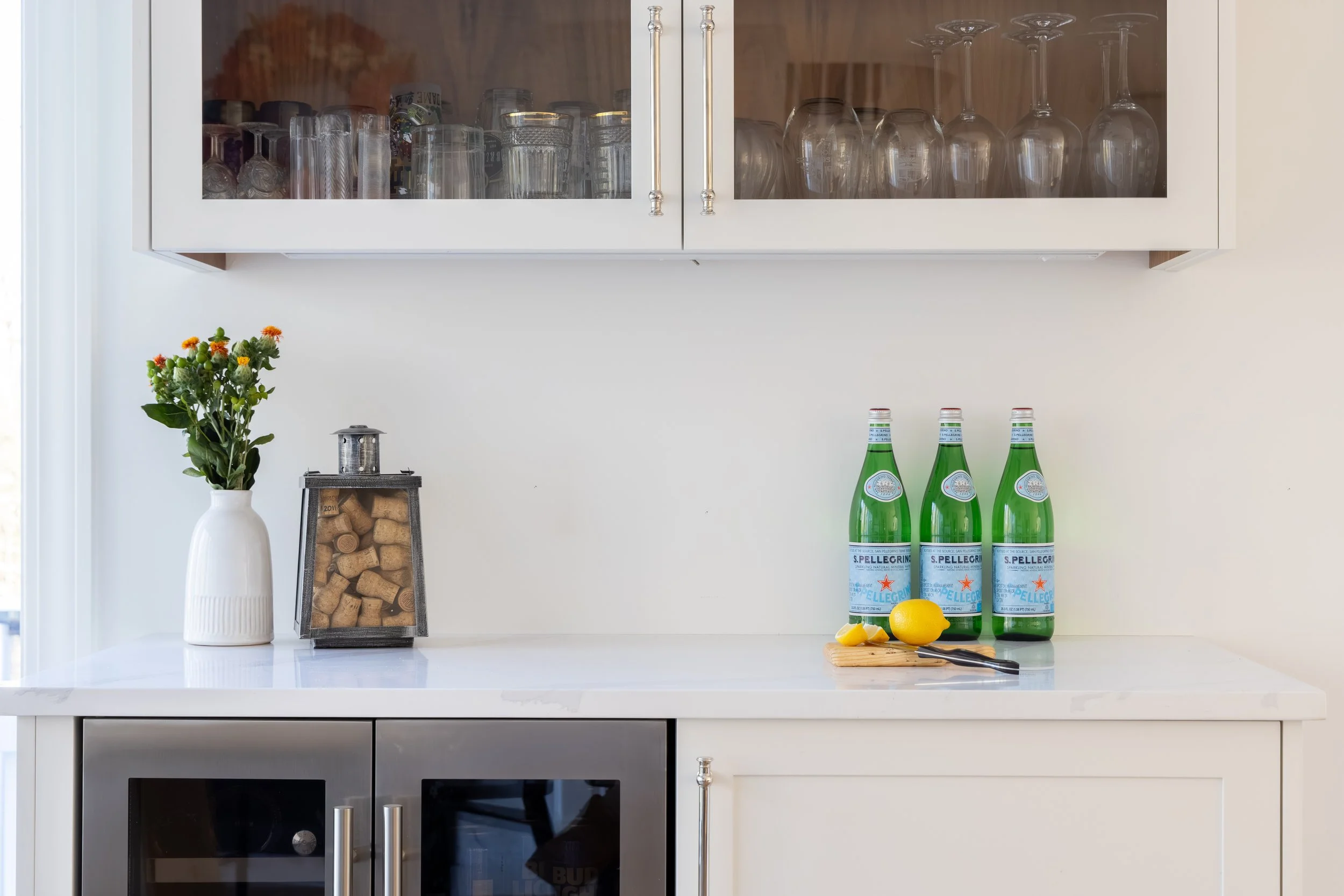Bright kitchen bar cabinet with glassware, a flower vase, wine cork display, and three San Pellegrino bottles with lemons on a cutting board