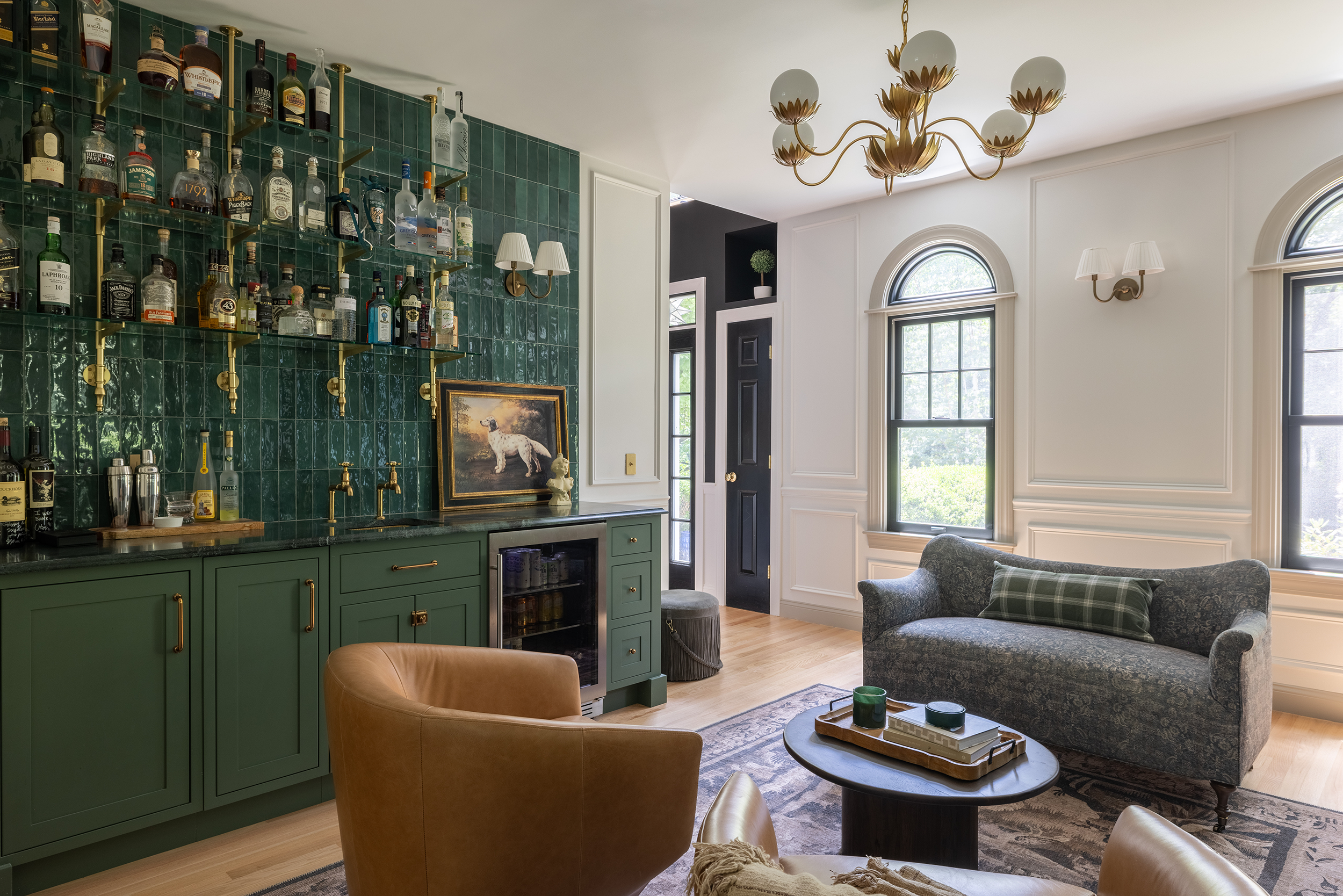 Elegant home bar and living room with green cabinetry, liquor shelves, arched windows, patterned sofa, and gold chandelier