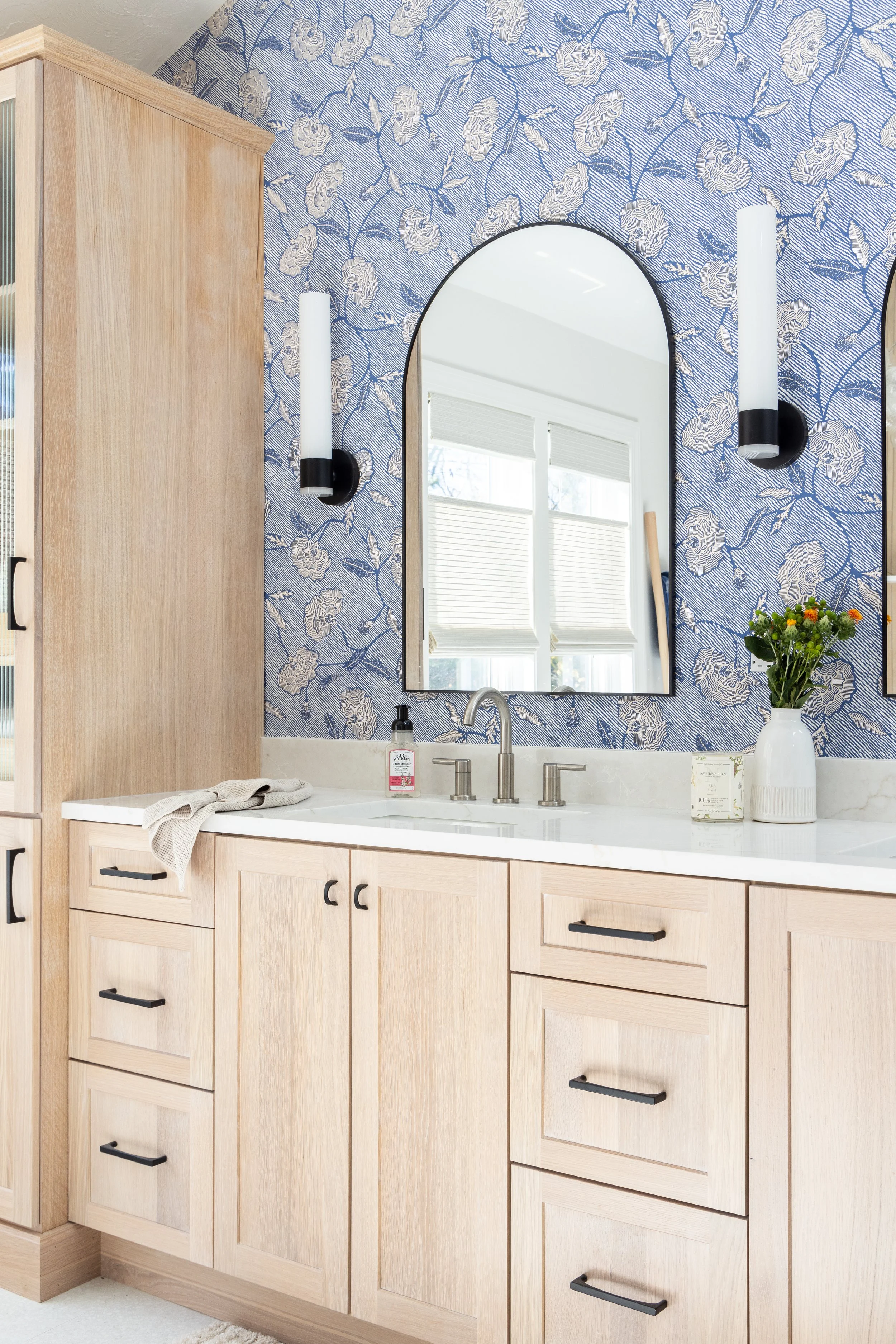 Modern bathroom vanity with light wood cabinets, white countertop, arched mirror, black sconces, and blue floral wallpaper