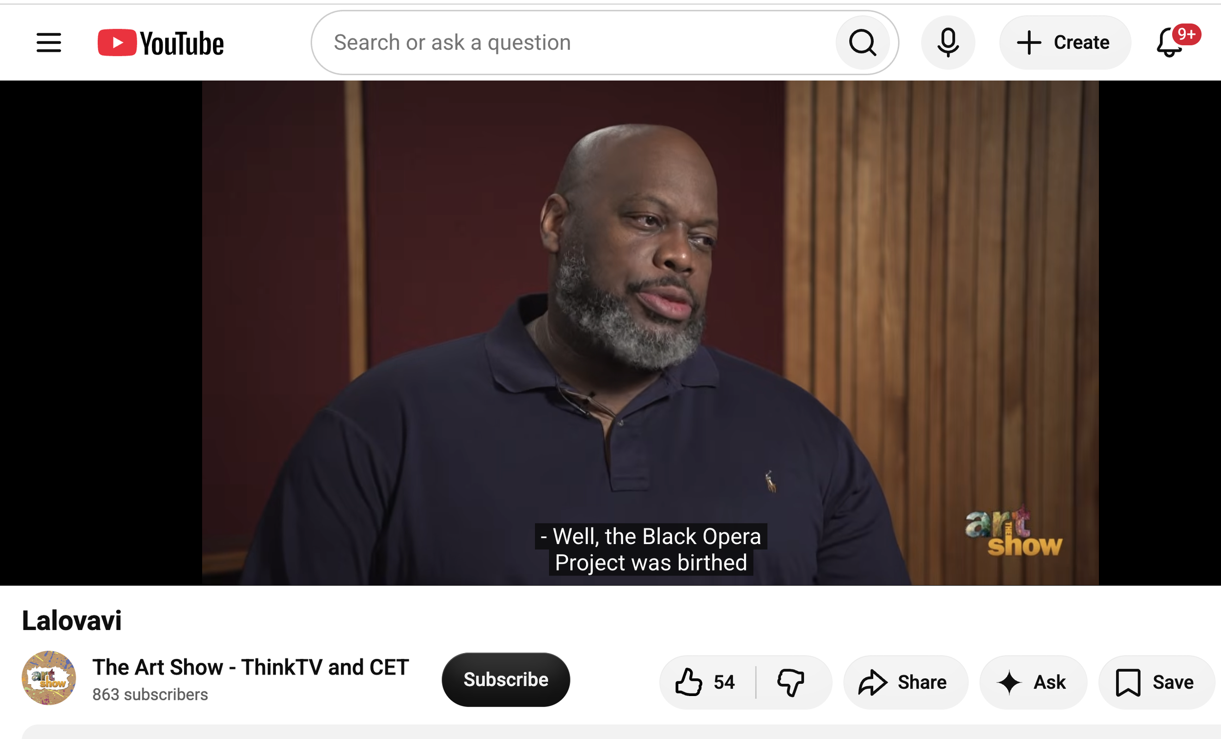A man with a bald head and beard wearing a navy blue polo shirt, sitting during an interview on a talk show set, with a wooden panel background. The subtitle reads: 'Well, the Black Opera Project was birthed'.