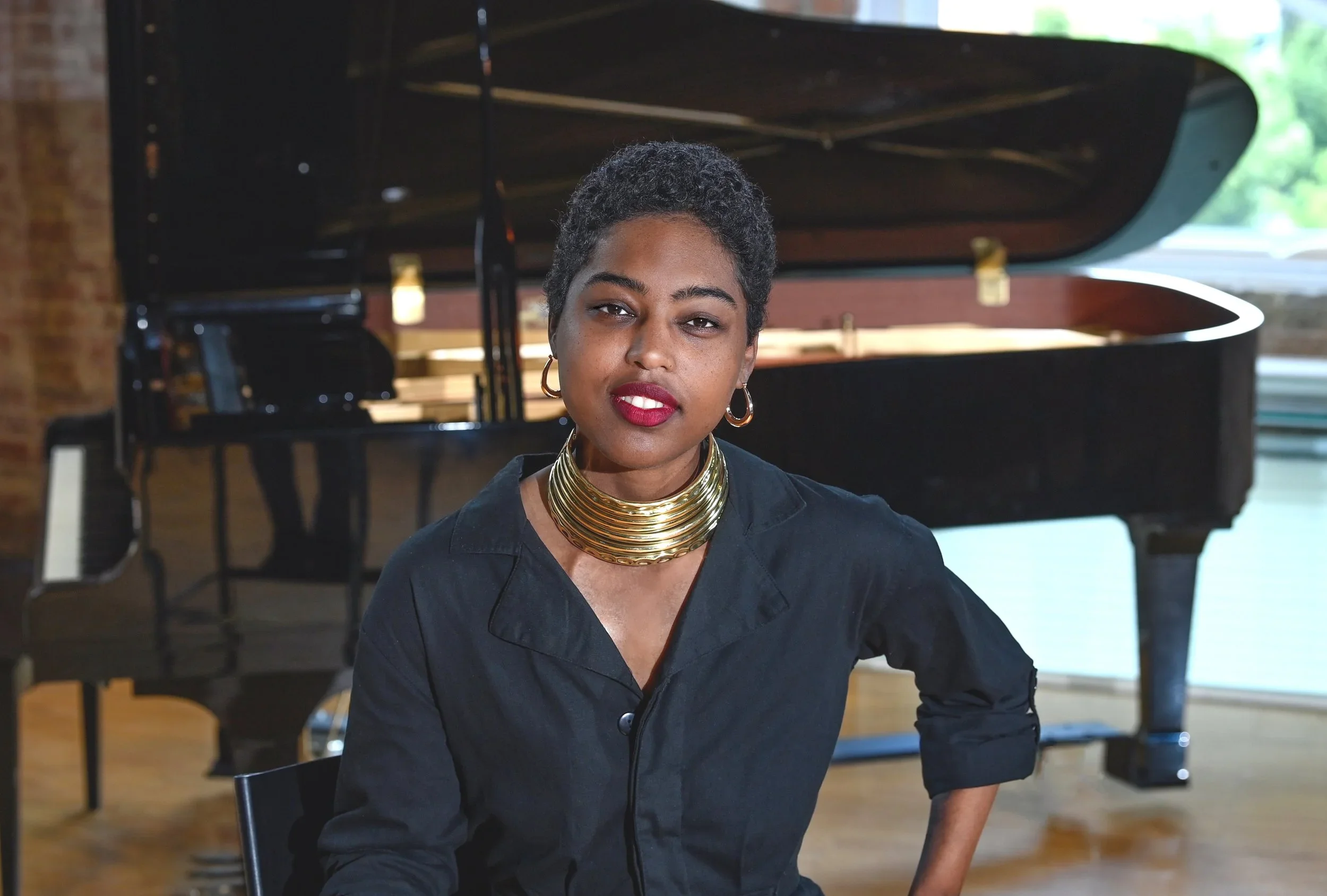 A woman with short curly black hair, wearing a black shirt, gold hoop earrings, and a layered gold necklace, sitting in front of a grand piano with a brick wall and large windows in the background.