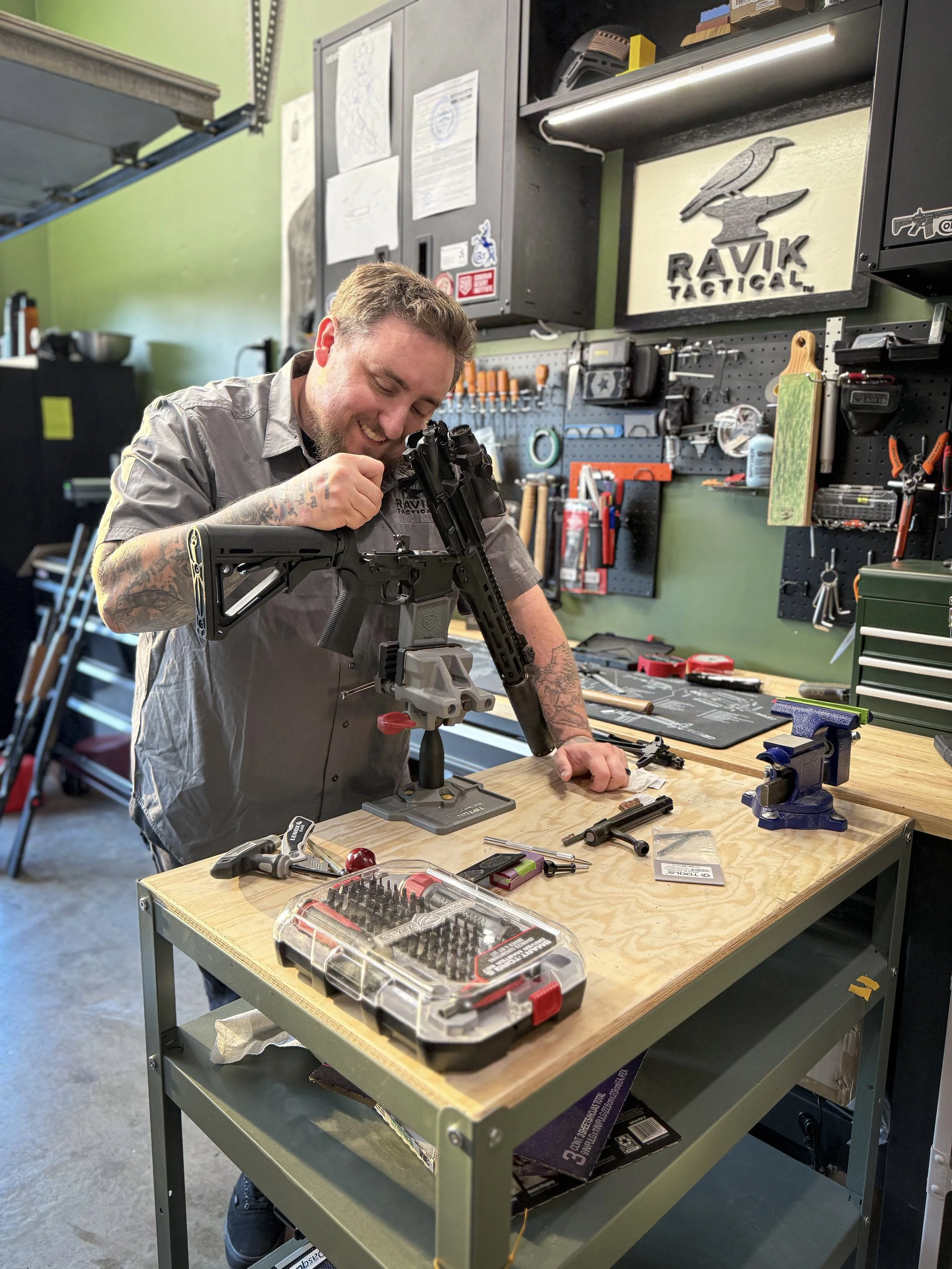 A man working on assembling or repairing a firearm at a workshop table, surrounded by tools, with a pegboard and wall-mounted storage in the background.