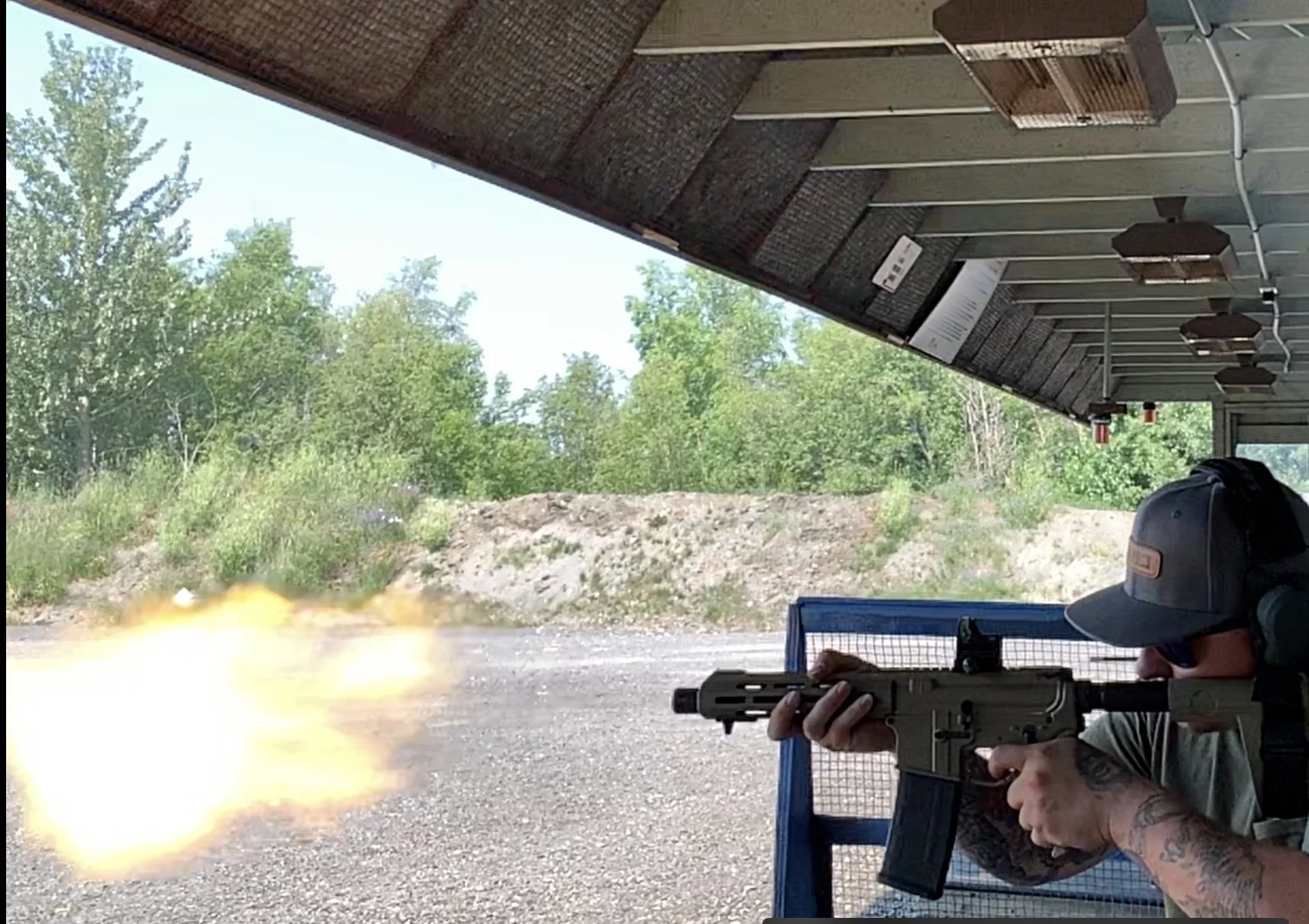 Person shooting a firearm at an outdoor shooting range with a visible muzzle blast, wearing a cap and tattoos on their arm.
