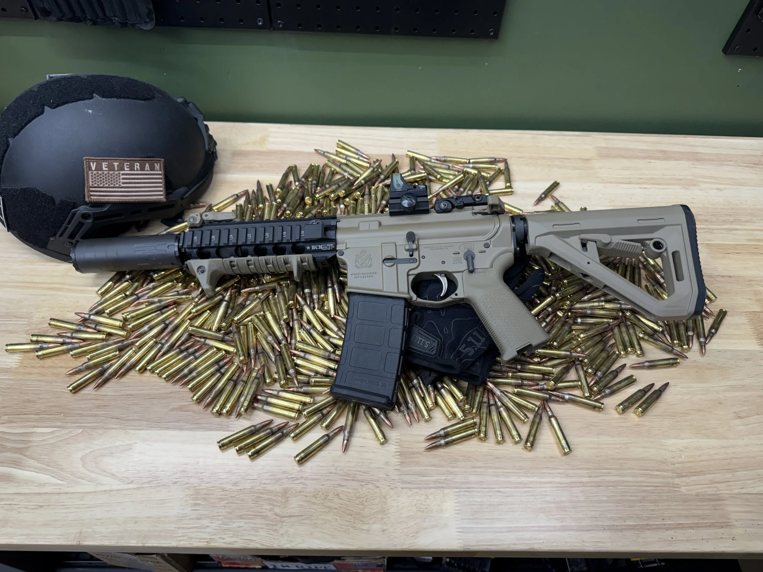 A firearm lying on a wooden surface surrounded by a large number of bullets, with a black helmet that has a 'Veteran' patch resting nearby.