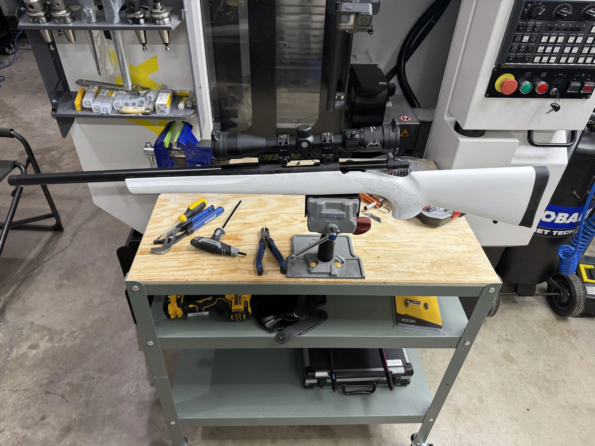 Workshop cart with tools and equipment, including a rifle scope mounted on a rifle, tools on the wooden work surface, drill, screwdriver, and pliers, a scale, safety glasses, and various machine parts.