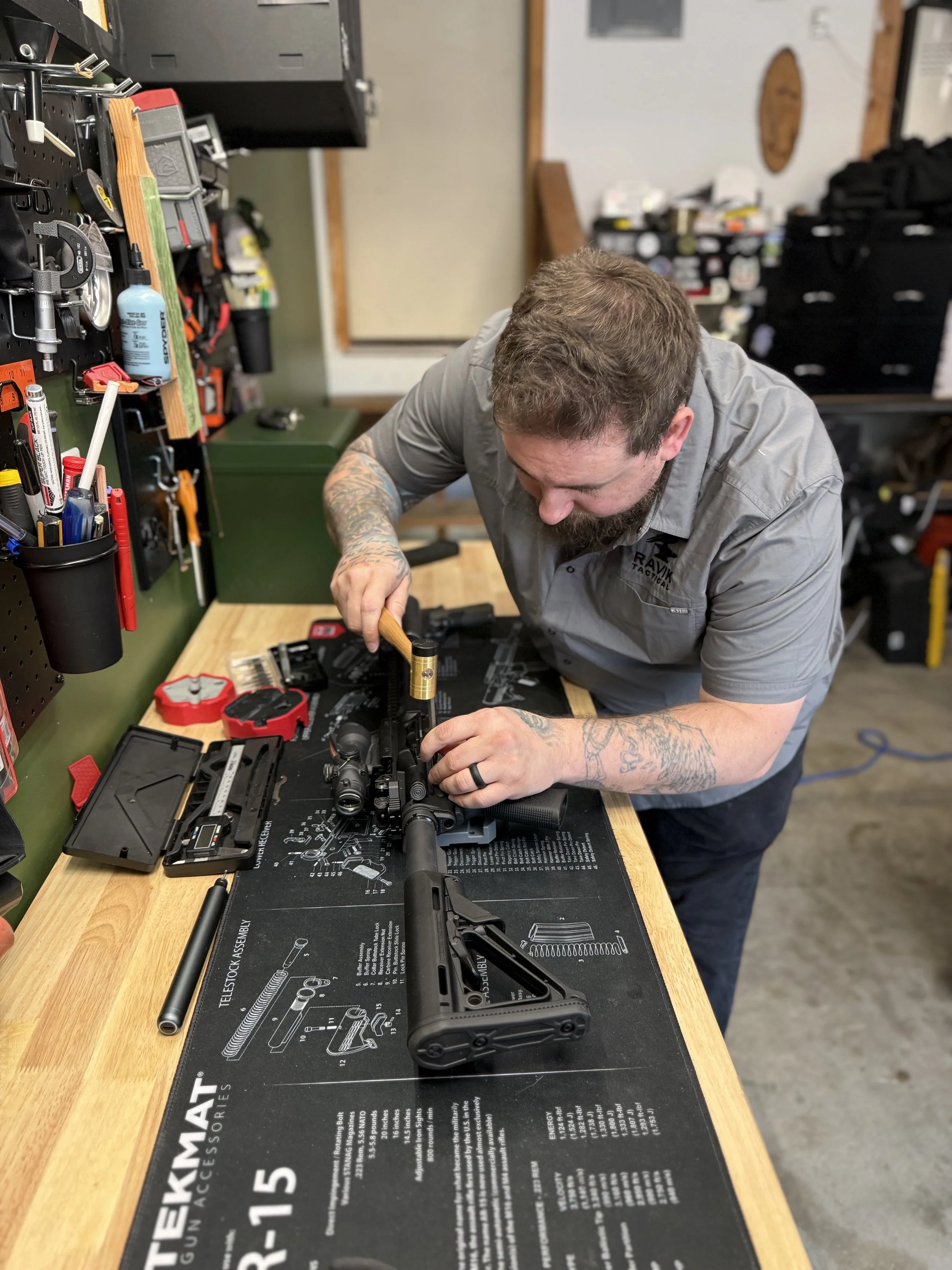 A man with tattoos on his right arm is working on assembling a rifle at a workbench in a workshop. The workbench has a diagram of firearm parts and tools scattered around.