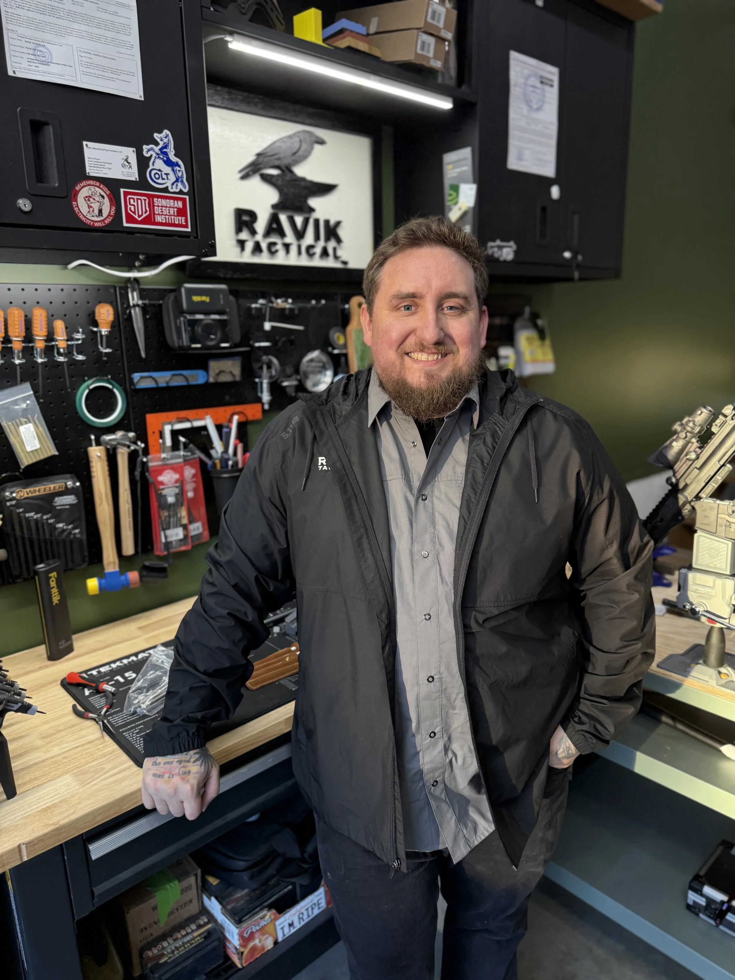 A man with a beard standing in a workshop, smiling, with his fist resting on a workbench. Behind him are organized tools, a sign that says 'RAVIK TACTICAL,' and a pegboard with various tools and supplies.