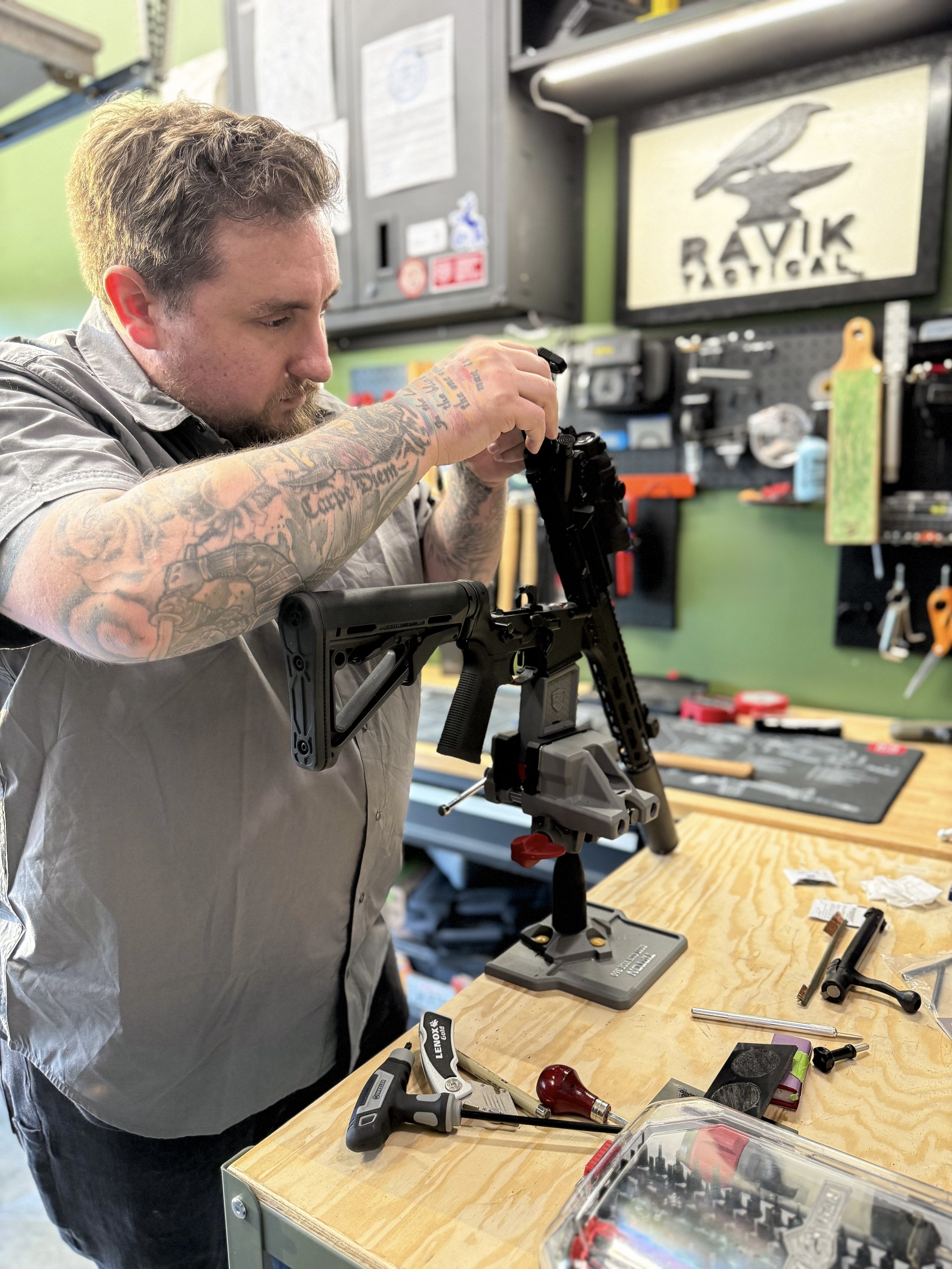 A man with tattoos on his arms working on assembling a firearm in a workshop. The workshop has various tools and parts on a wooden workbench and a shelf with a sign and equipment in the background.