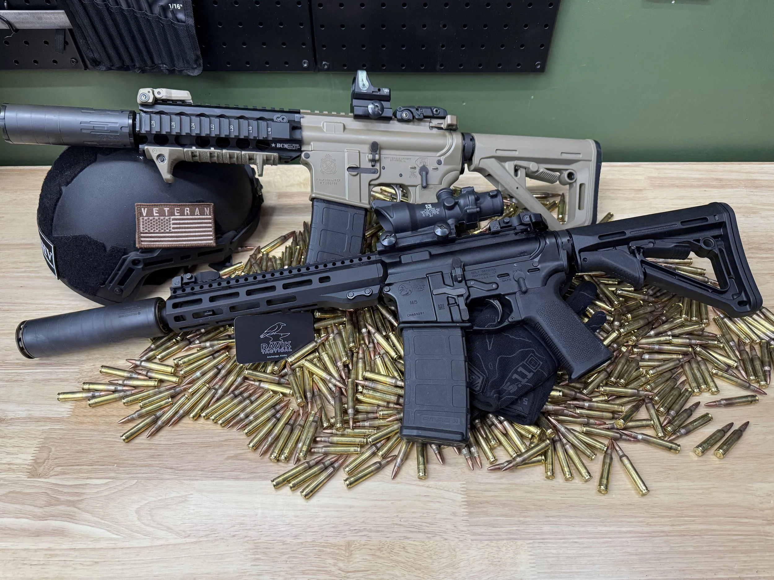 A firearm with a scope and suppressor, lying on a table alongside numerous brass cartridge casings, a tactical helmet with a veteran patch, and a magazine.