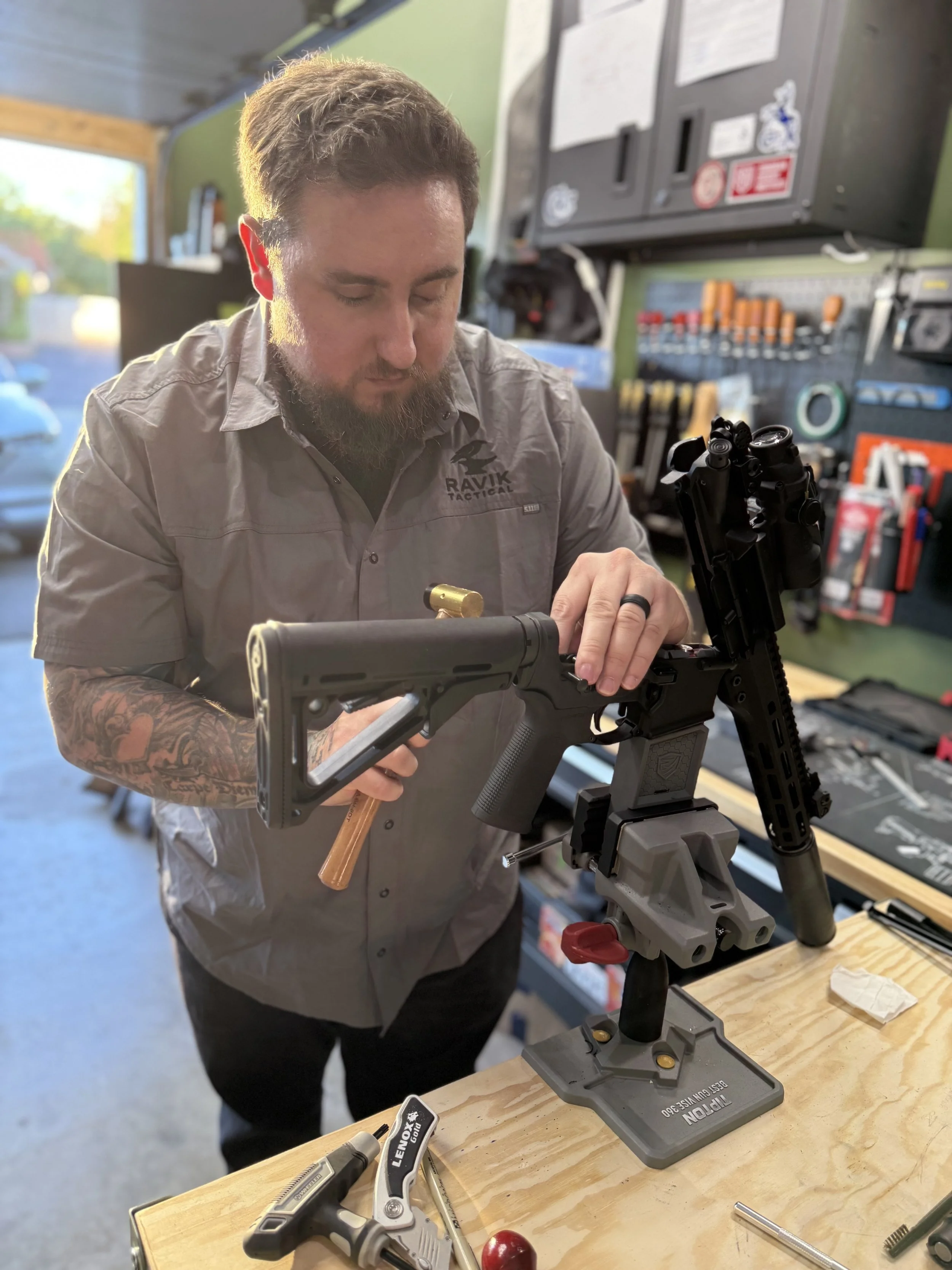 A man working on assembling a firearm using tools in a workshop. He is wearing a gray shirt with a logo that says 'RAVIK TACTICAL' and has tattoos on his arms. The workbench has various tools and supplies.