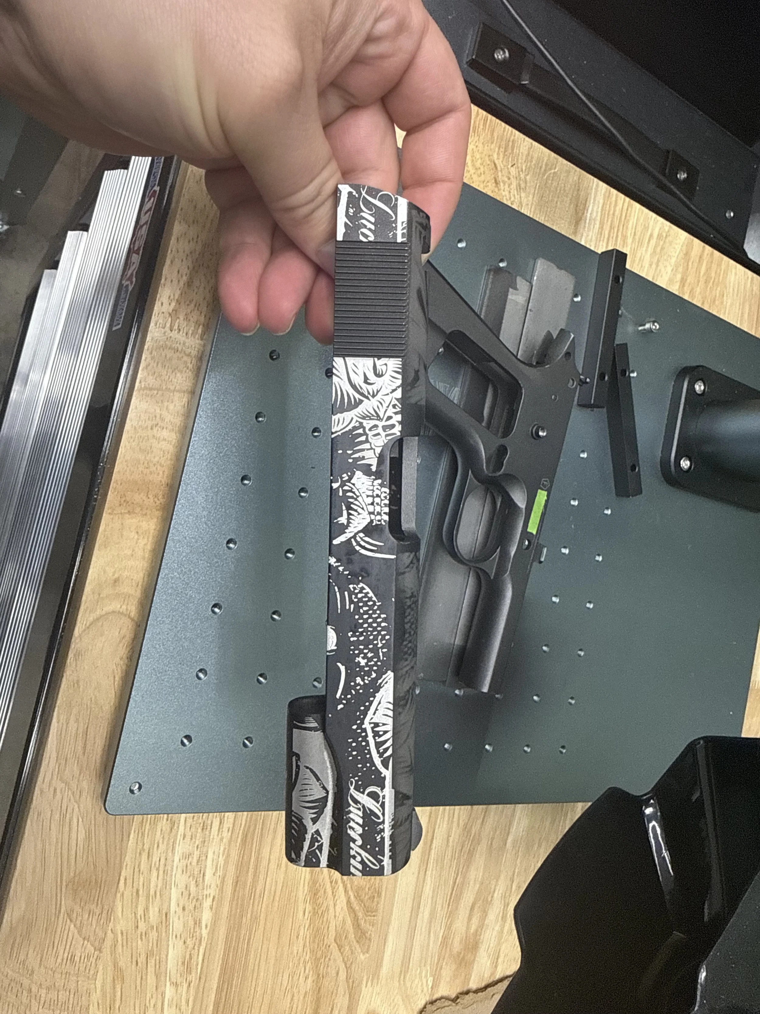 A handgun with a black and white graphic design resting on a gray circuit board or work surface, with a wooden table edge and black chair in the background.