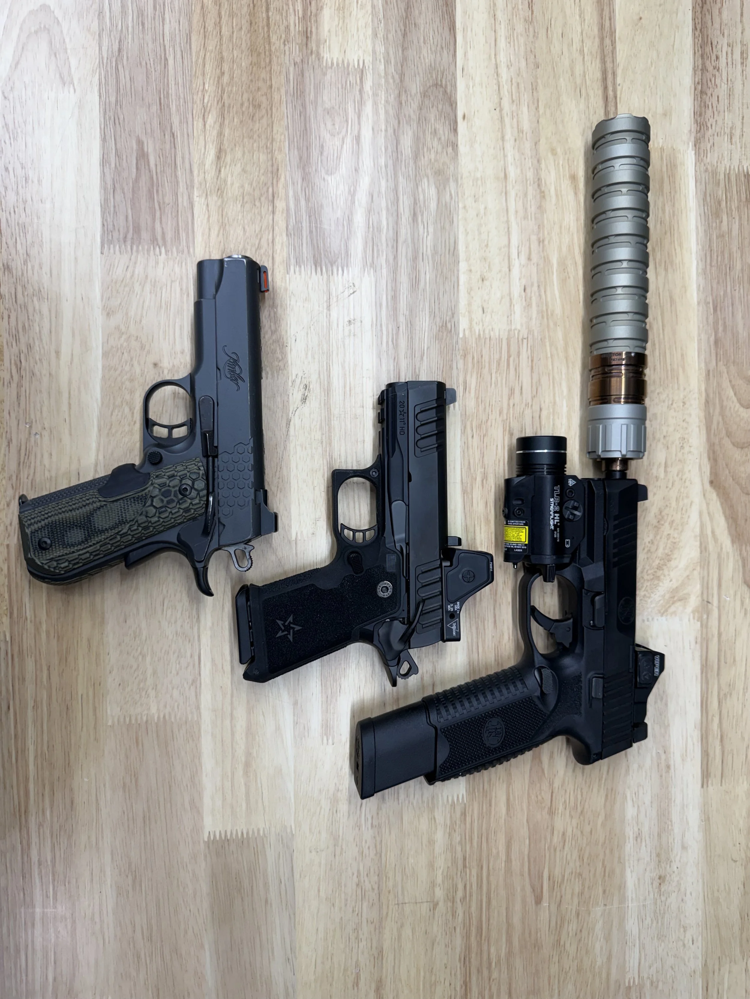 Array of three firearms including two handguns and a rifle with tactical accessories, placed on a wooden surface.
