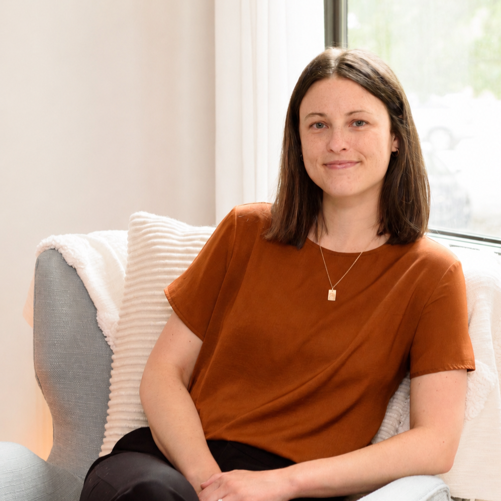 Kate Murphy, psychotherapist, with shoulder-length brown hair, wearing a copper colored shirt and gold necklace, sitting on a light blue armchair next to a window with a beige cushion behind her in the chair