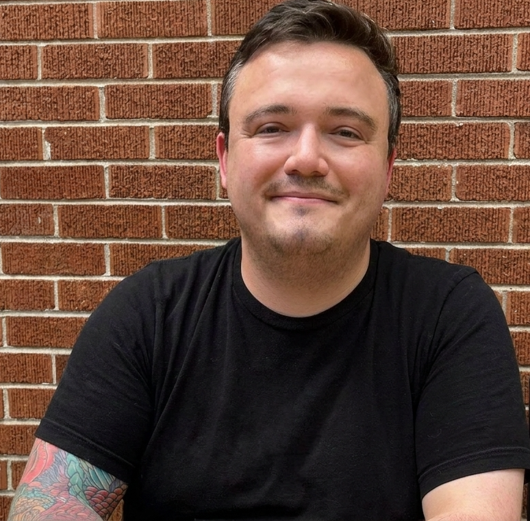 A white man with a black shirt, smiling, colorful tattoos on his right arm in front of a red-brick wall - aCT Licensed Counselor in Evanston, Illinois - Serving Residents in Illinois and Indiana