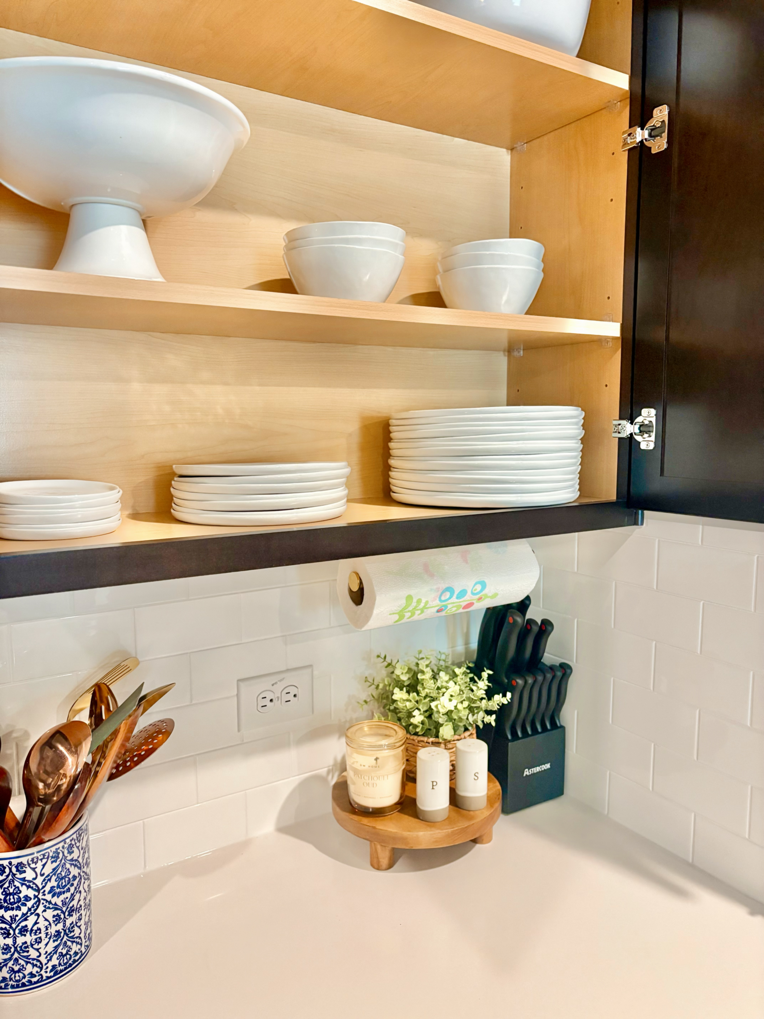 Kitchen cabinet open with white dishes and bowls on shelves, paper towel, knives, utensils, candles, and decorative plants on the countertop.
