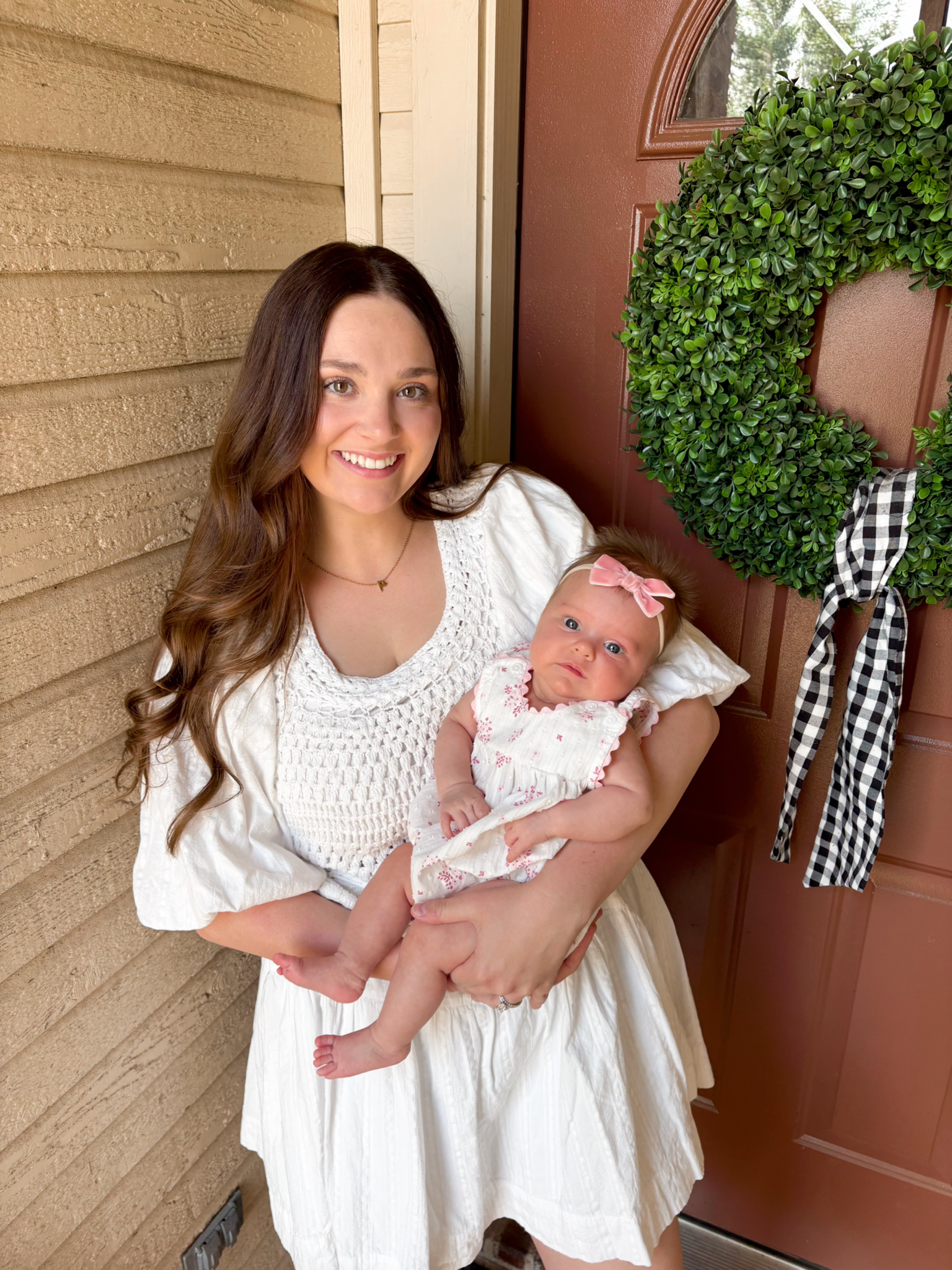 A woman with long brown hair smiling and holding a baby girl with a pink bow in her hair, standing near a door with a wreath and a wooden wall.