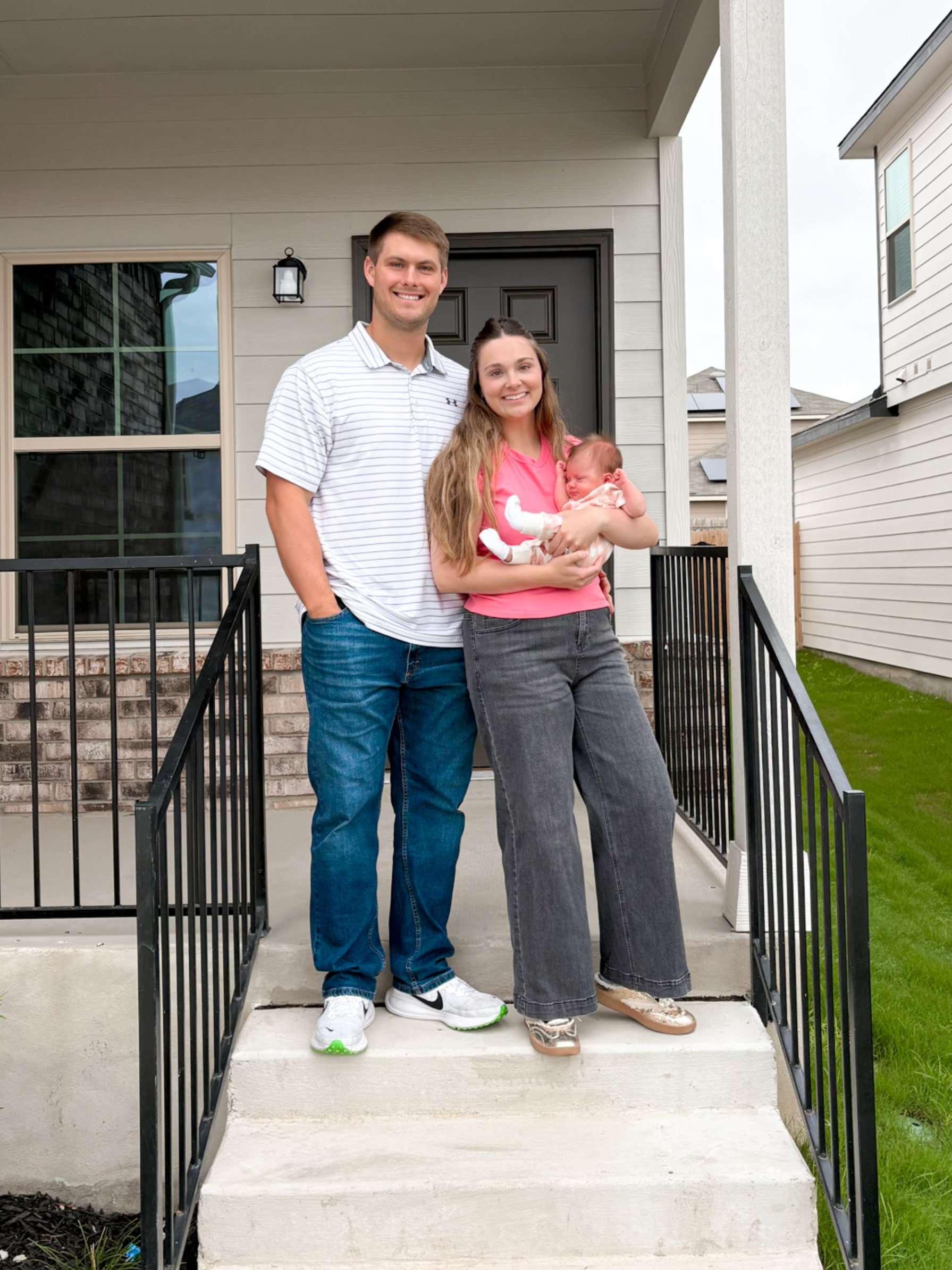 A smiling couple standing on the front porch of their house, holding their newborn baby. The man is wearing a white striped shirt and jeans, and the woman is dressed in a pink shirt and gray jeans, with the baby in her arms.