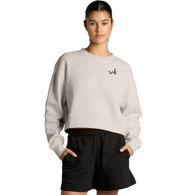 Young woman with dark hair in a ponytail wearing a beige cropped sweatshirt with black writing and black shorts, standing against a plain white background.