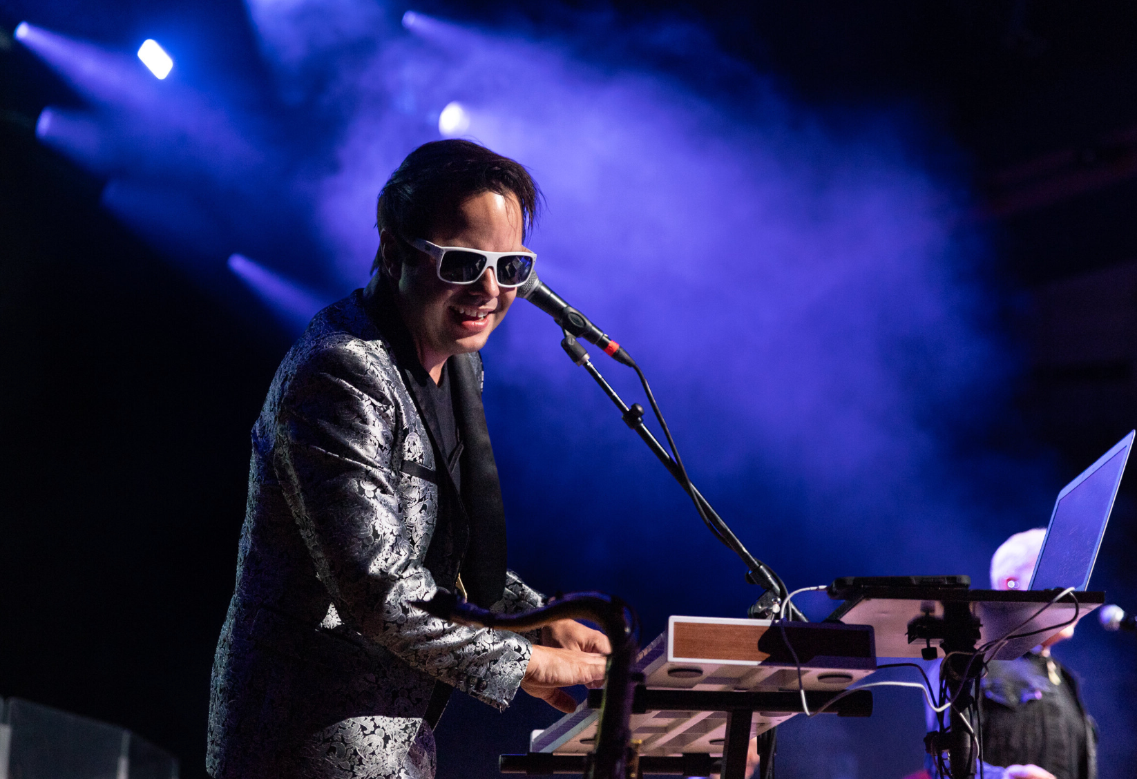 A male musician wearing sunglasses and a floral patterned jacket playing a keyboard on stage with purple and blue stage lights.