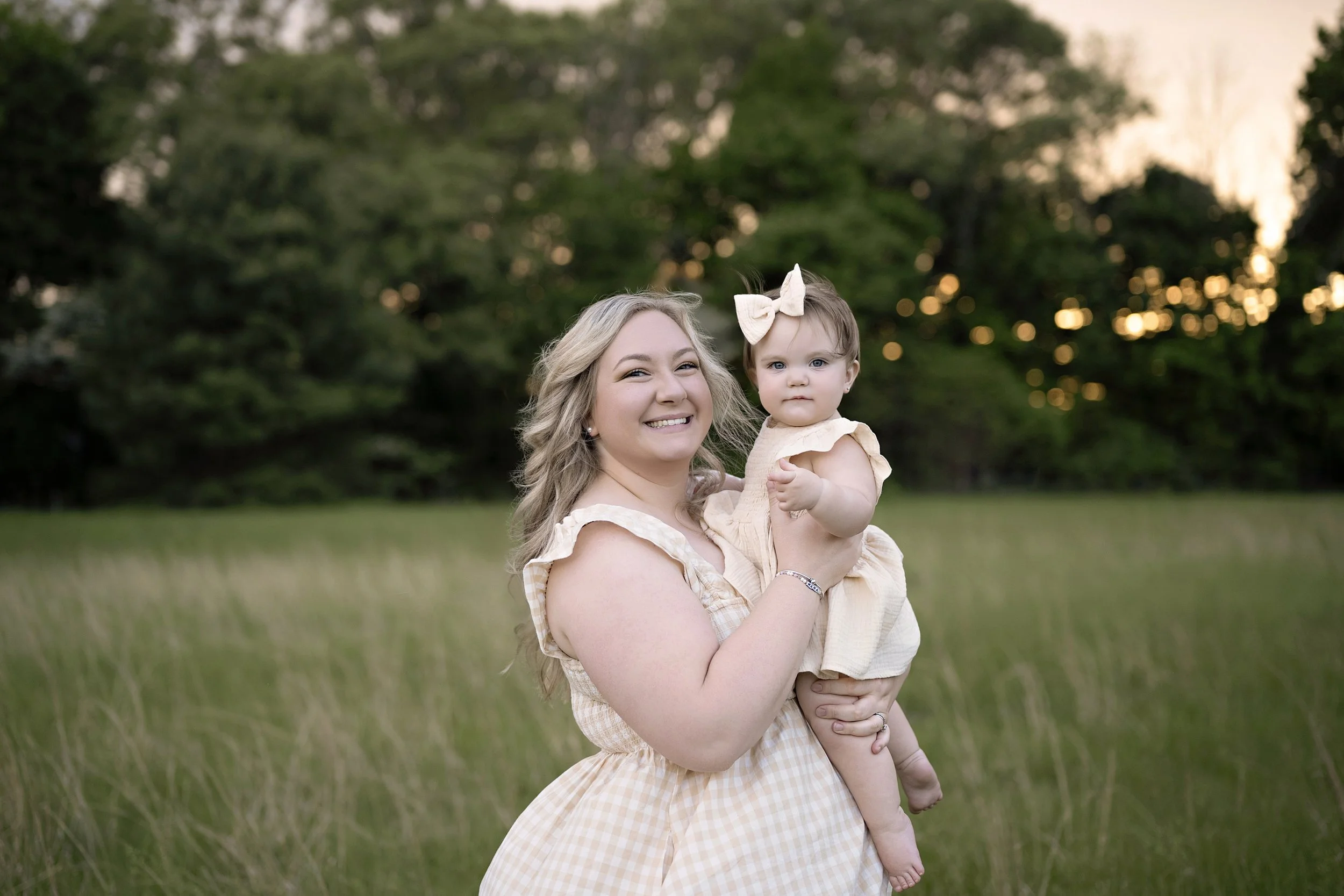 A woman with blonde hair holding a young girl with a beige bow in her hair outdoors with trees and a sunset in the background.