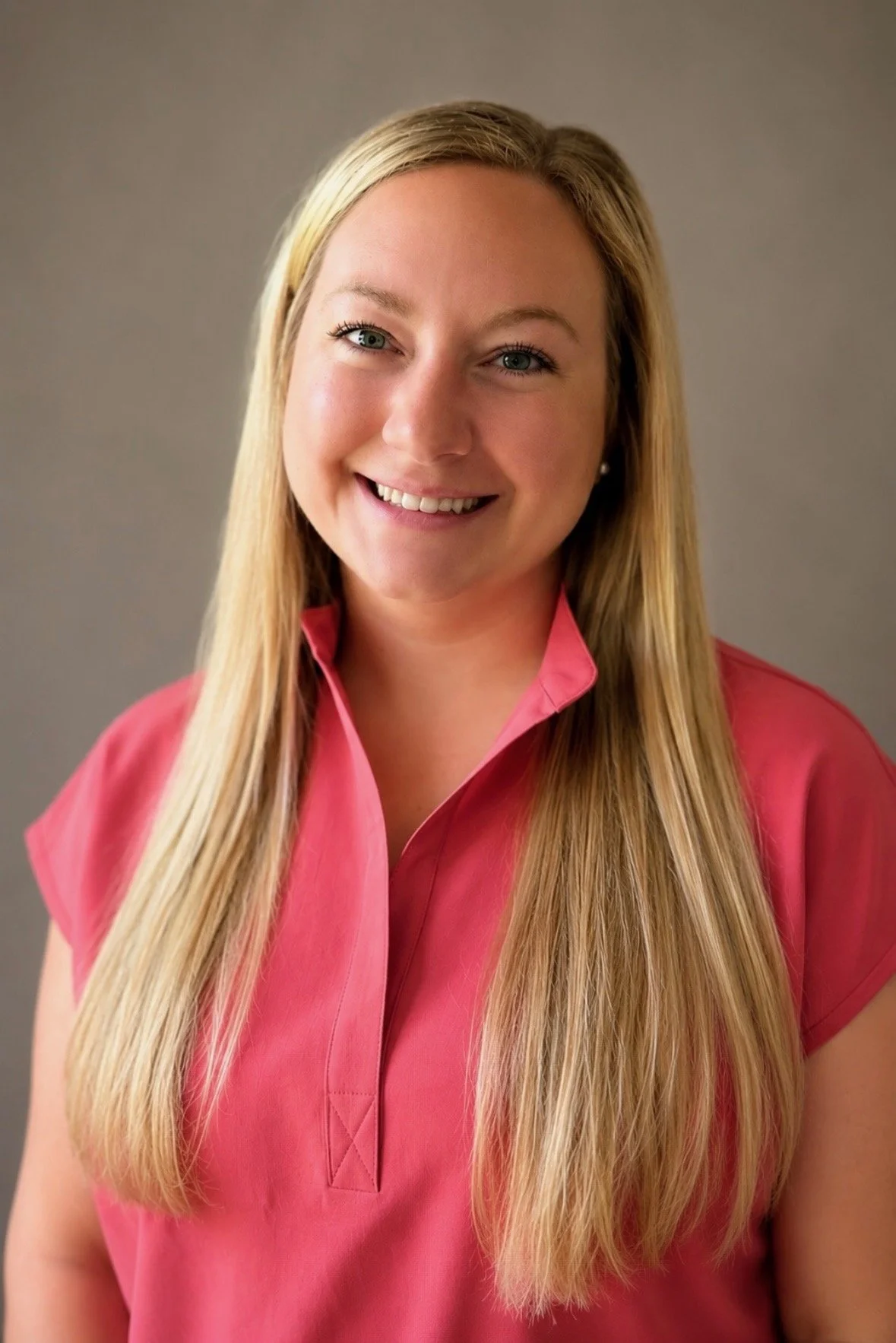 A woman with long blonde hair wearing a pink collared shirt, smiling against a plain gray background.