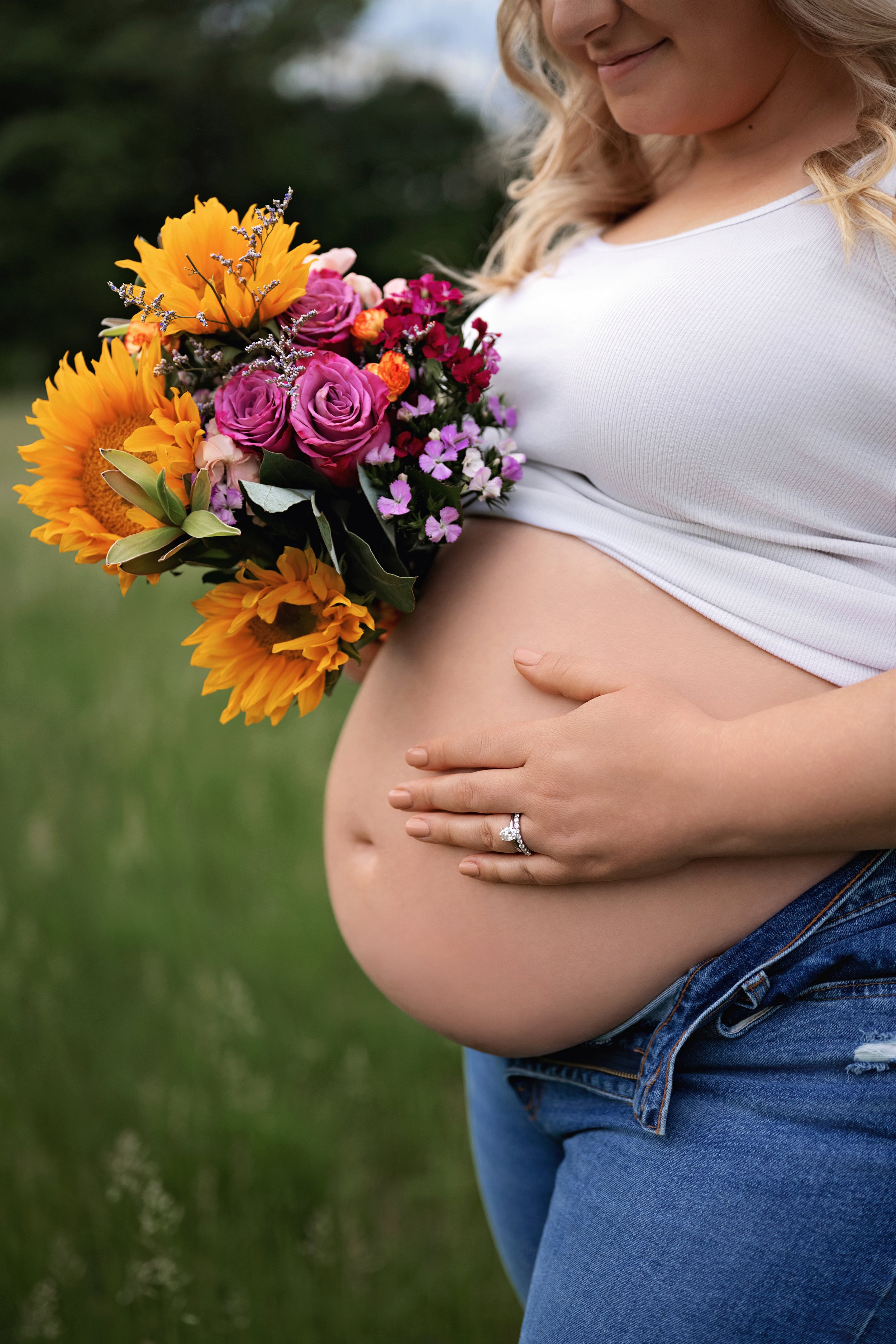 Close-up of a pregnant woman holding a colorful bouquet of flowers on her belly, wearing a white top and blue jeans outdoors.