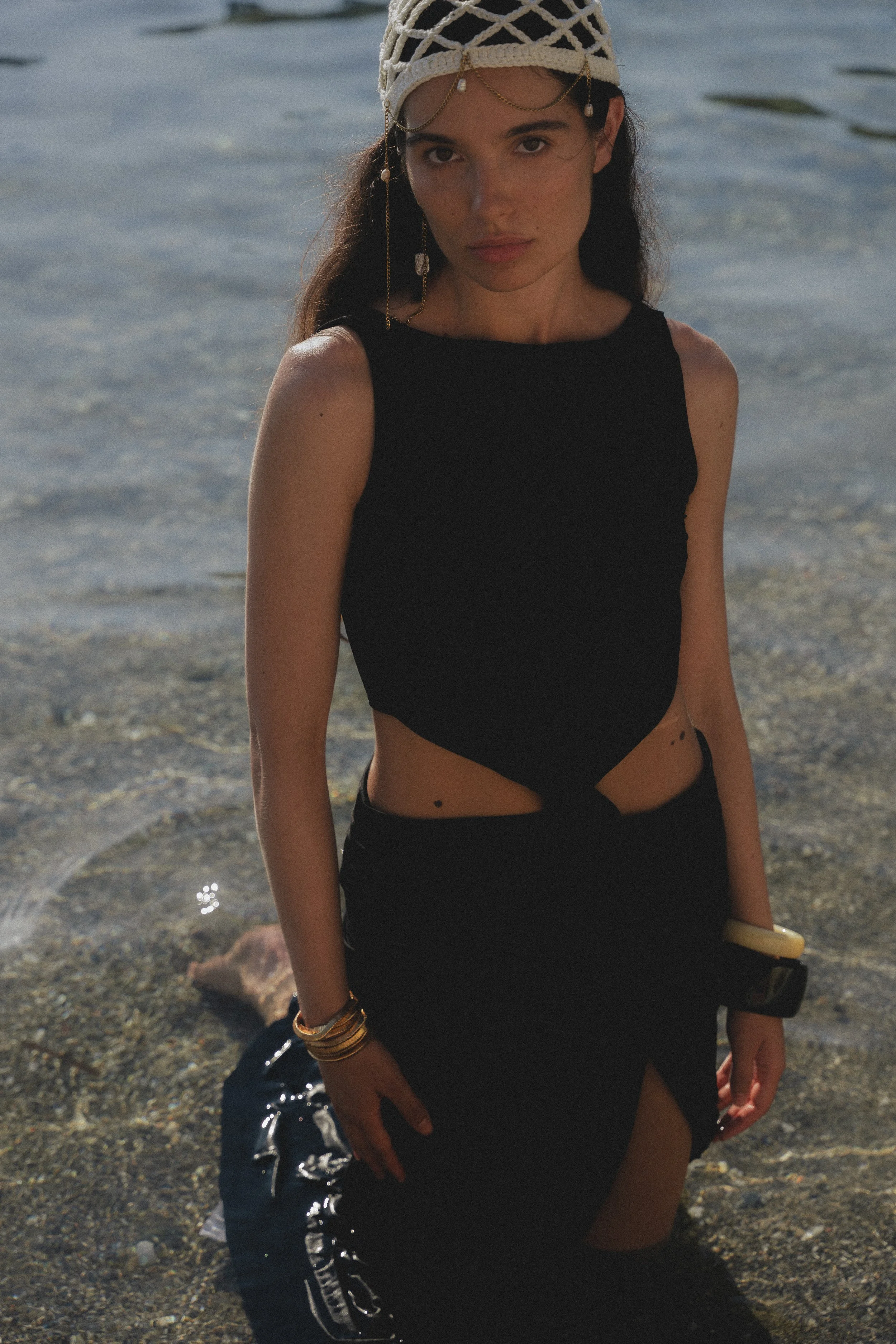 A woman with long dark hair standing by the water on a beach, wearing a black sleeveless top, black skirt, jewelry including a headpiece and bracelets, with the ocean and rocks in the background.